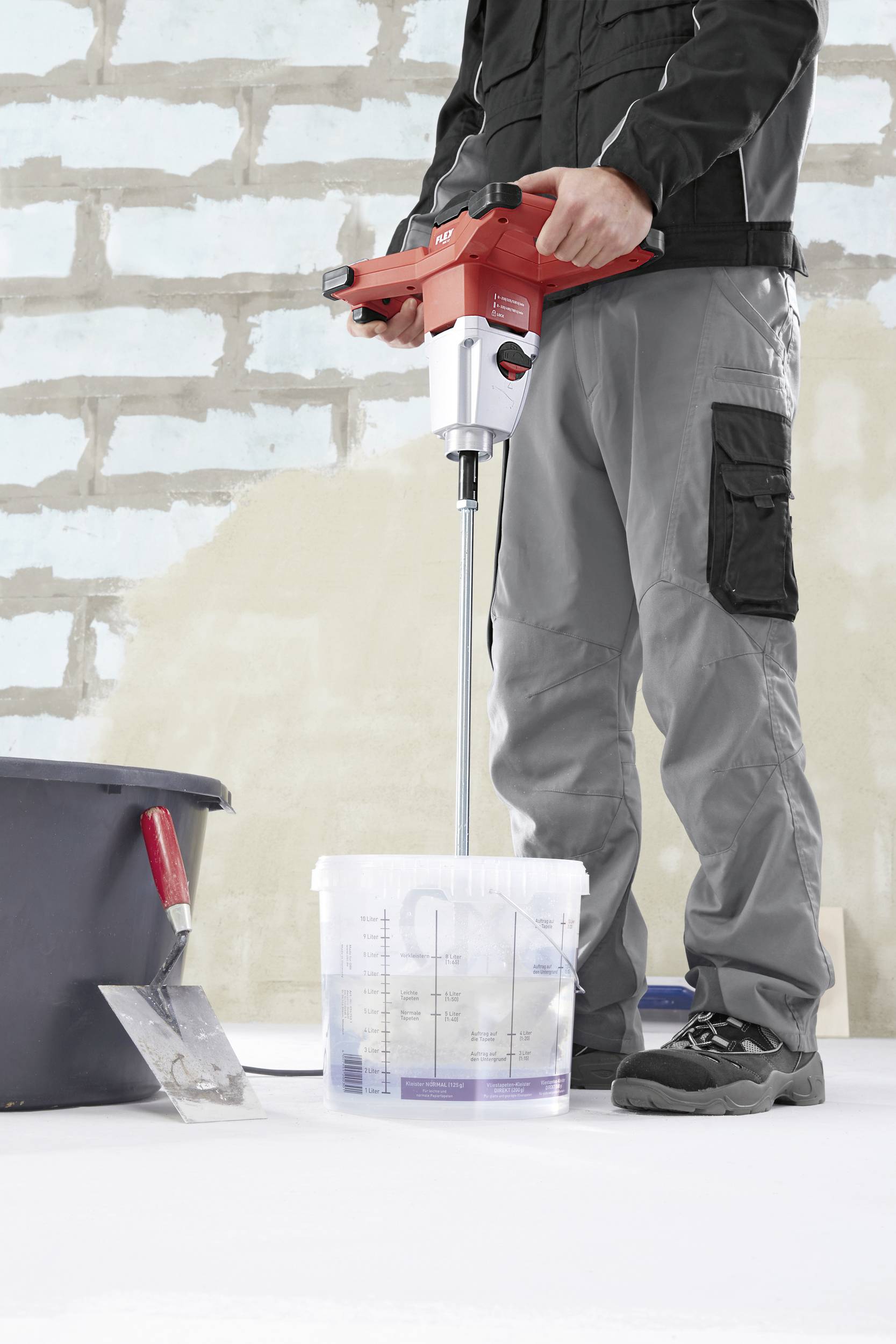 Osoba miesza płyn elektrycznym mikserem w wiadrze, obok leży chochla na podłodze budowy.