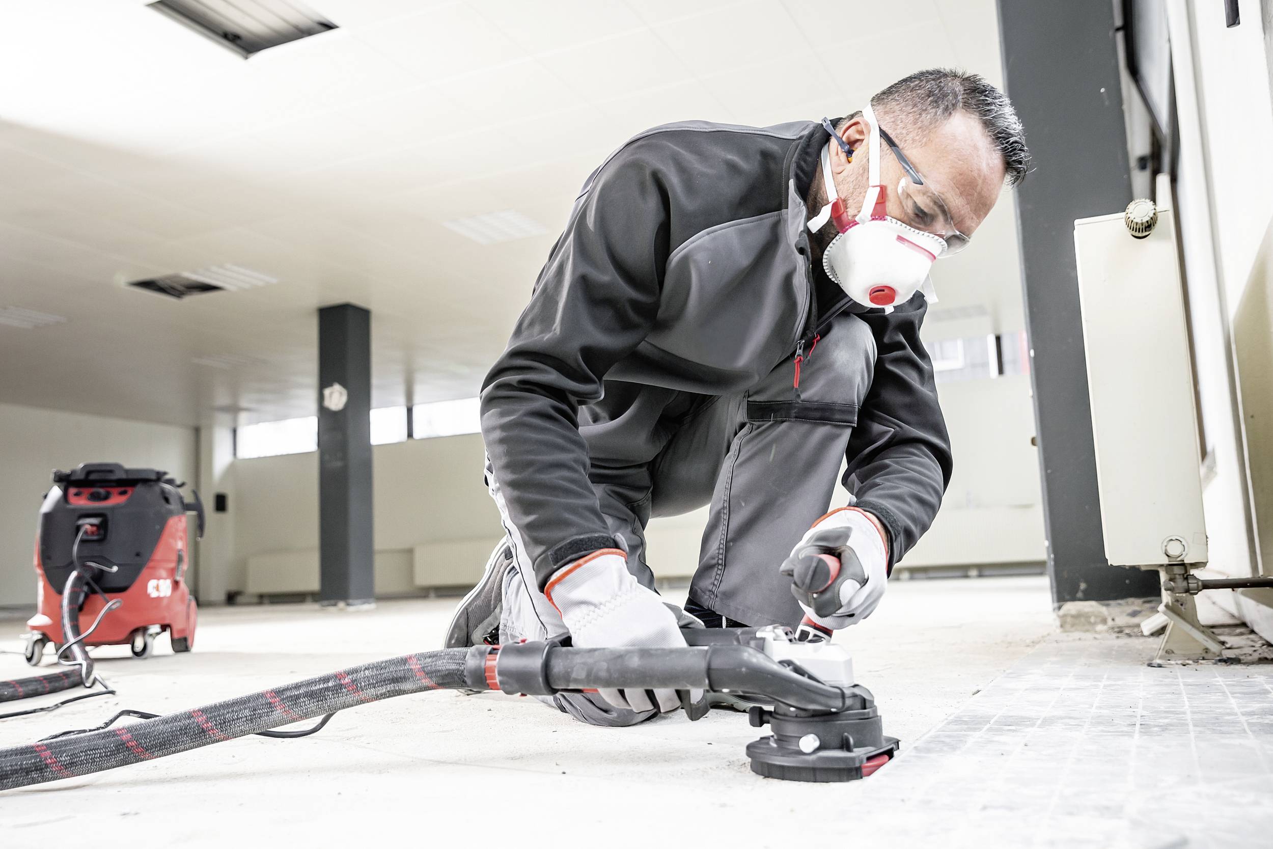 Robotnik w odzieży ochronnej i masce pracuje z narzędziem do prac podłogowych. W tle widoczny jest przemysłowy odkurzacz.