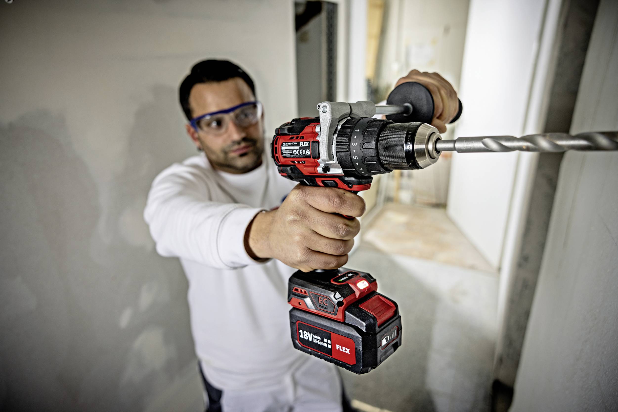Mężczyzna w okularach ochronnych używa mocnego akumulatorowego wiertarko-wkrętarki, aby wywierić otwór w ścianie. Pracuje z pełnym skupieniem w niedokończonym pomieszczeniu.