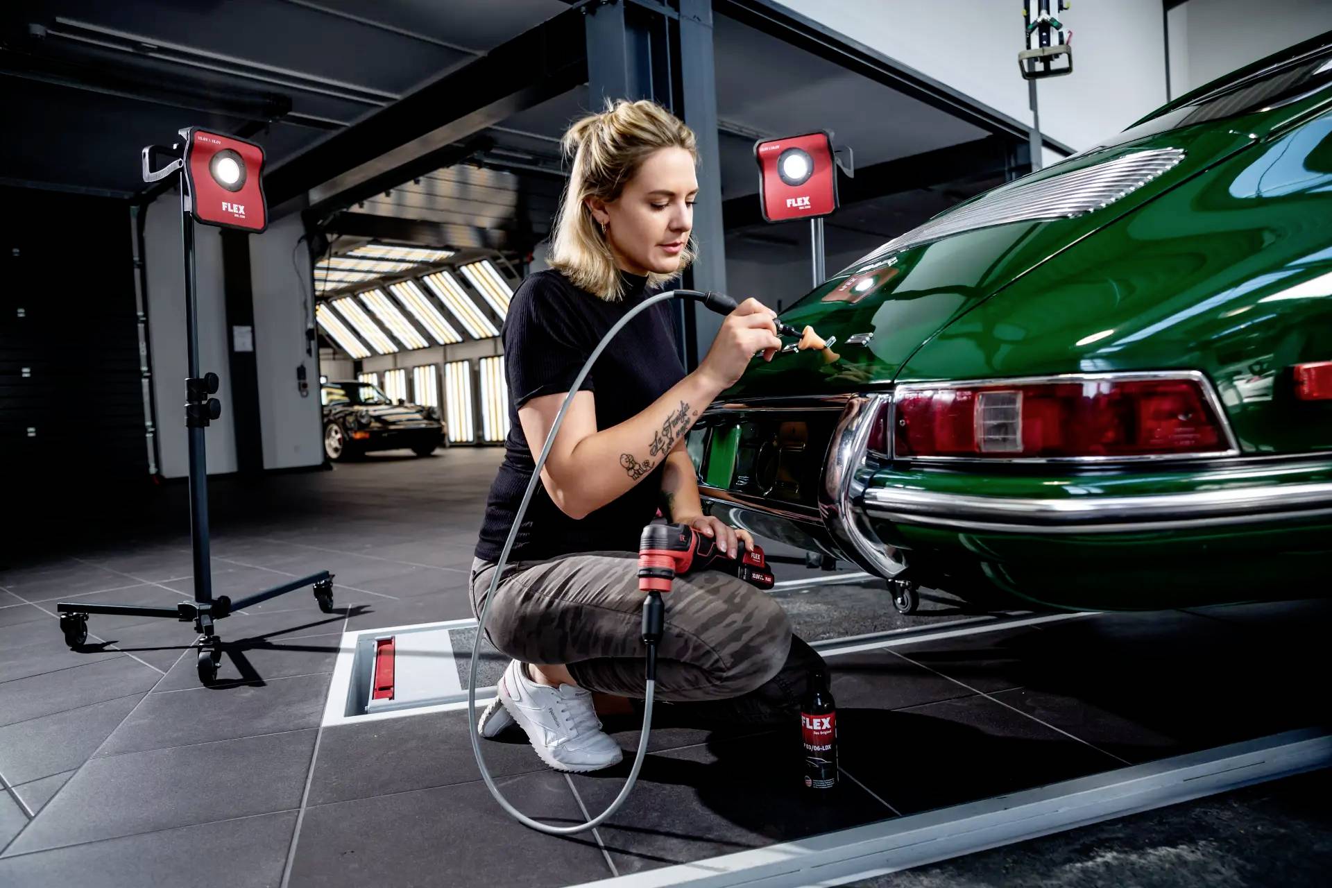 Kobieta poleruje tylną część zielonego zabytkowego samochodu w warsztacie z metalowym, połyskliwym otoczeniem. Używa specjalistycznych narzędzi.
