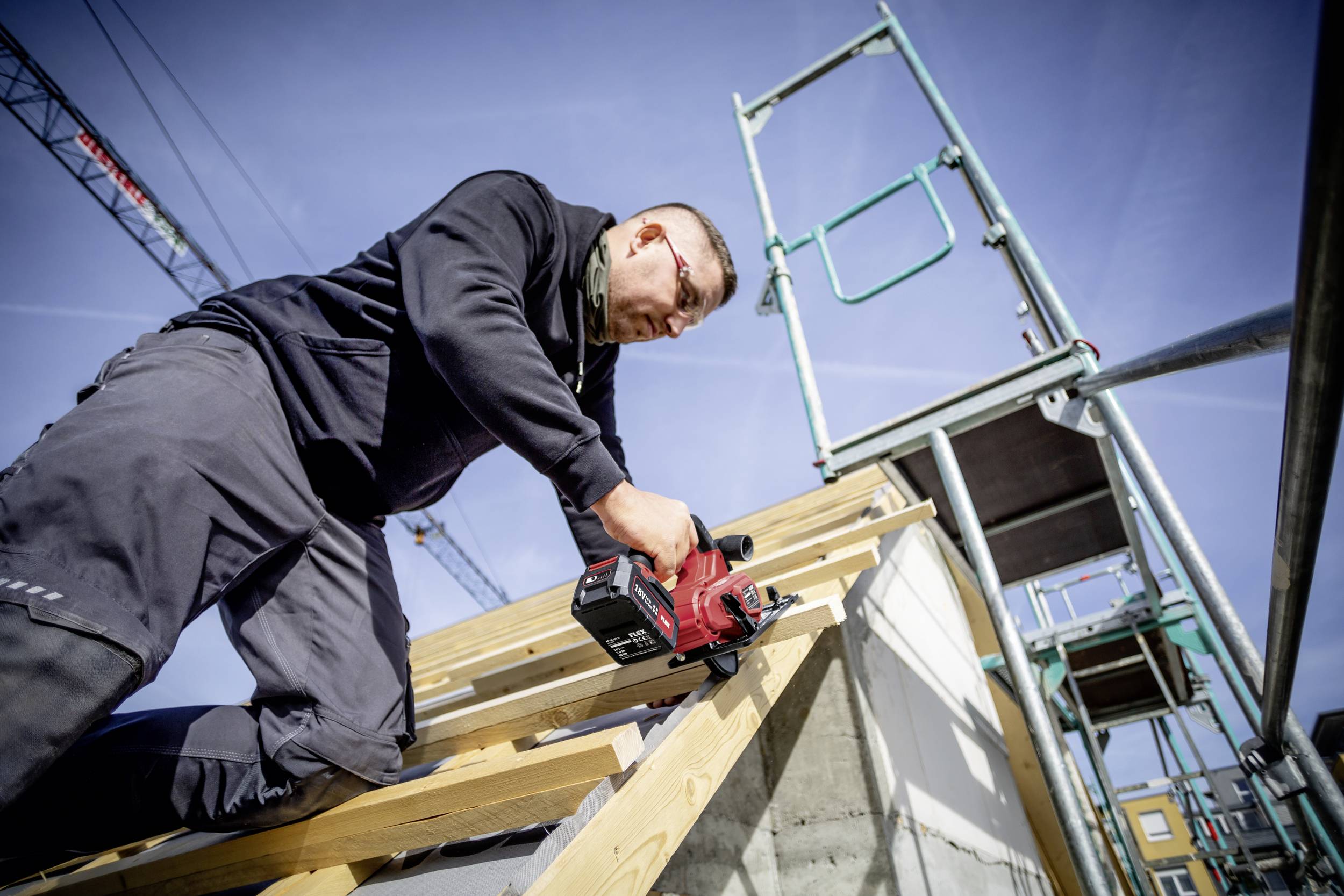 Robotnik na budowie używa elektronarzędzia do mocowania desek. W tle widoczne są rusztowania.