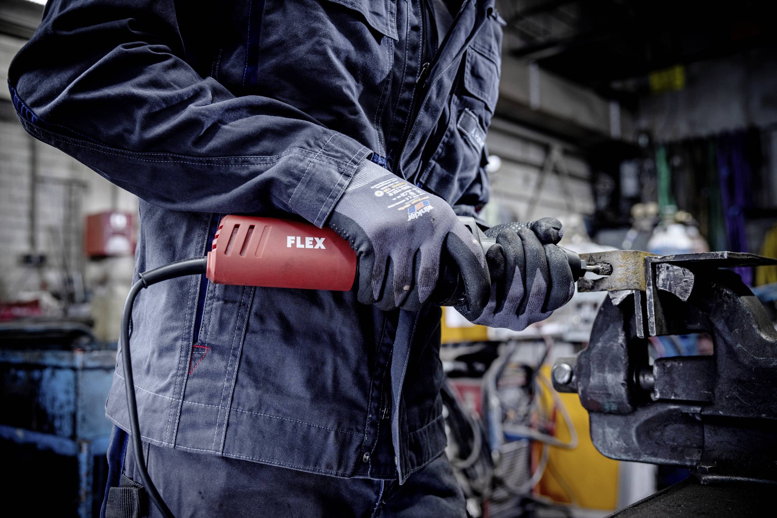 Osoba ubrana w strój roboczy trzyma w ręku elektronarzędzie, prawdopodobnie szlifierkę, w środowisku warsztatu.