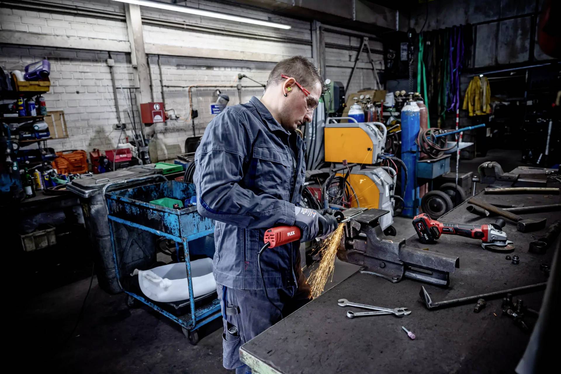 Mężczyzna szlifuje metal w warsztacie, wokół niego tryskają iskry. W tle widoczne są narzędzia i maszyny. Ma na sobie okulary ochronne.