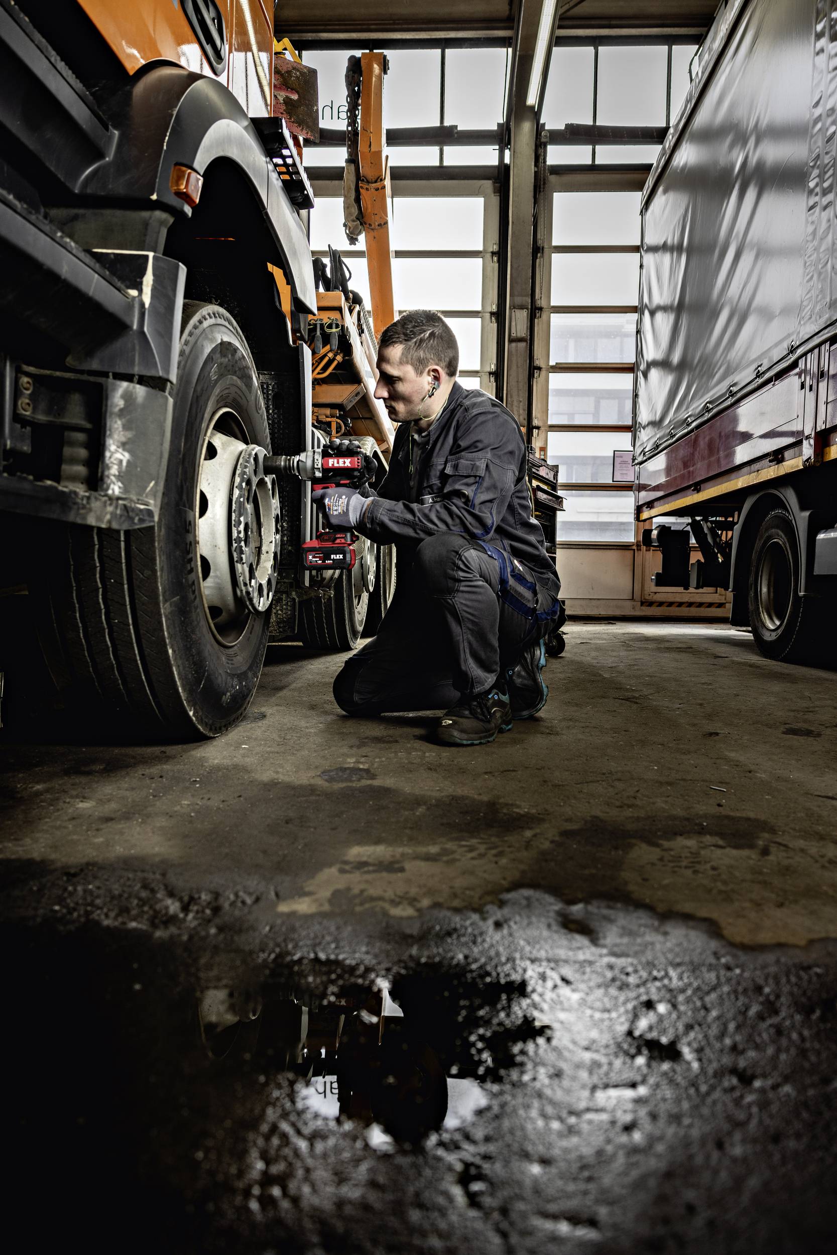 Mechanik naprawia przednie koło ciężarówki w warsztacie. W tle widać kolejne samochody ciężarowe.