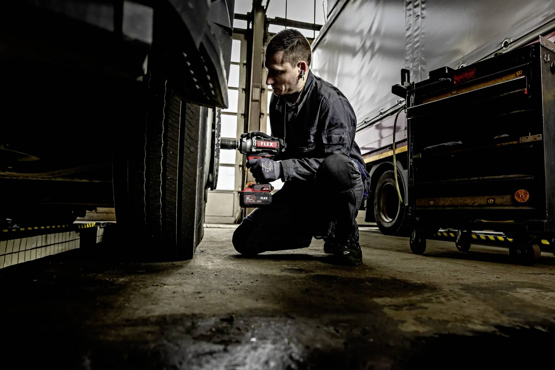 Mechanik pochyla się obok opony ciężarówki w warsztacie i używa elektrycznej wiertarki. W tle znajduje się wózek z narzędziami.