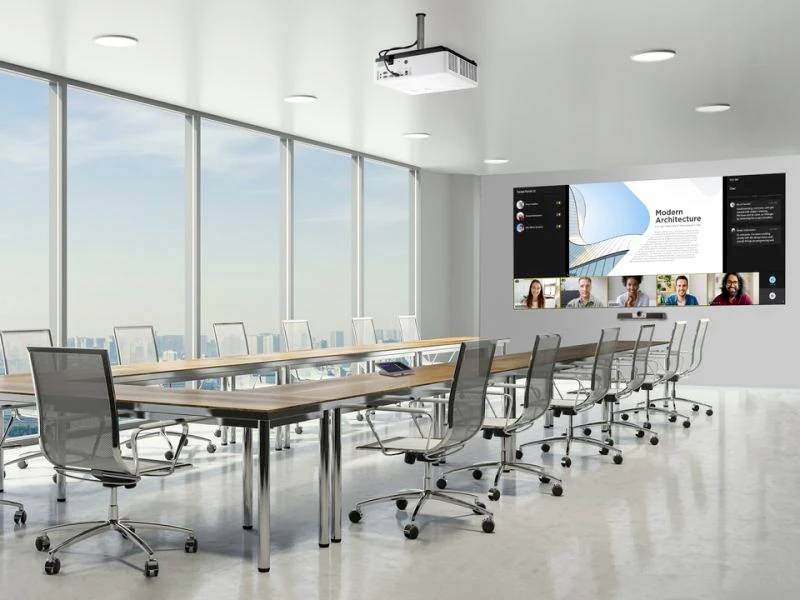 Duży pokój konferencyjny z prostokątnym stołem i kilkoma fotelami biurowymi. Na ścianie duży ekran wyświetlający wideokonferencję.