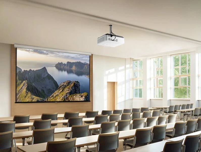 Pusty pokój lekcyjny z projekcją krajobrazu górskiego z morzem na ekranie. Duże okna wpuszczają światło dzienne.
