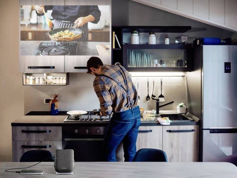 Mężczyzna gotuje w nowoczesnej kuchni, podczas gdy na ekranie wyświetlane jest wideo kulinarne. Ma na sobie koszulę w kratę i szelki.