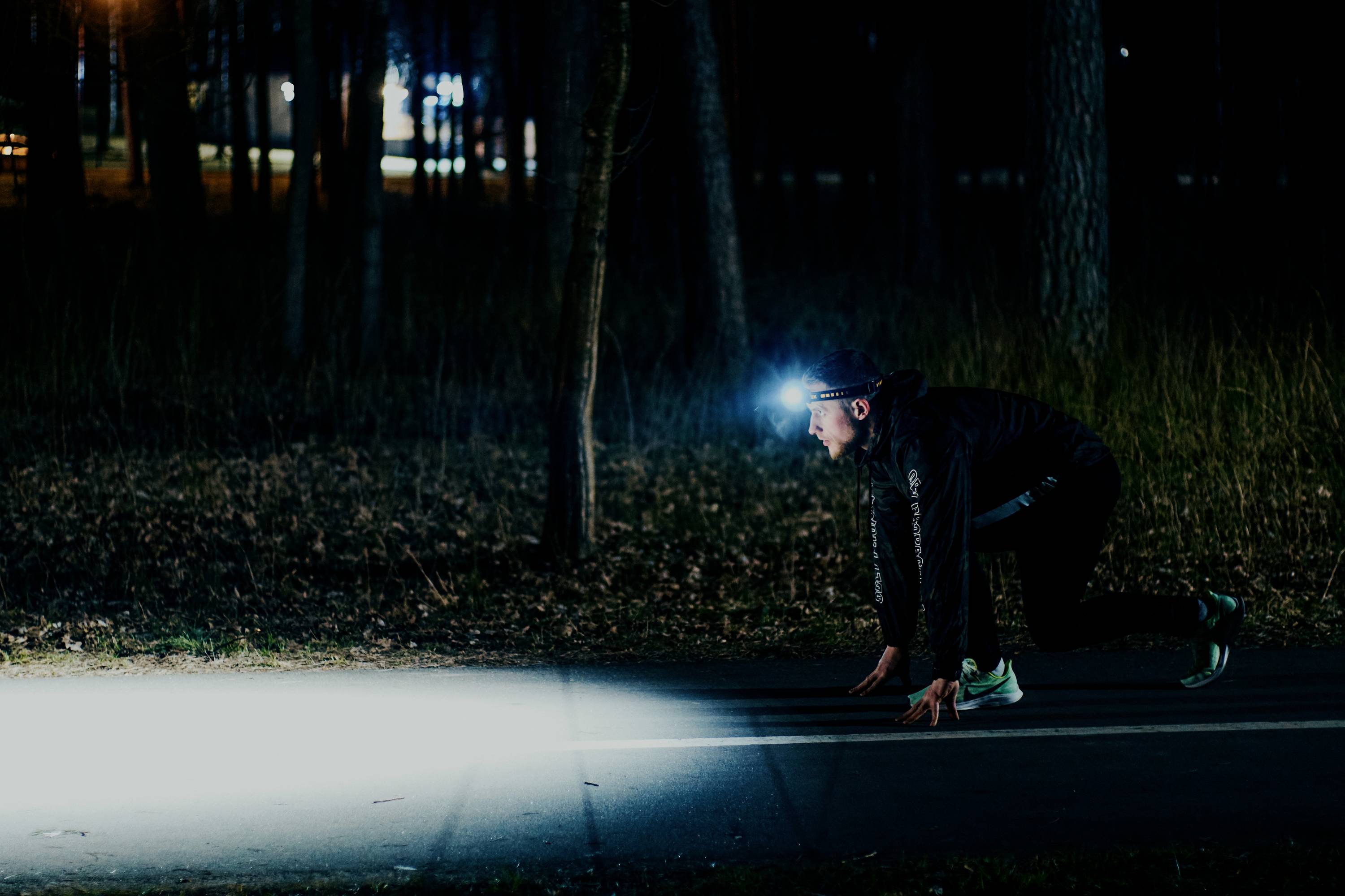 Osoba w stroju biegowym stoi nocą w pozycji startowej na oświetlonej ścieżce w lesie. Ma na głowie czołówkę.
