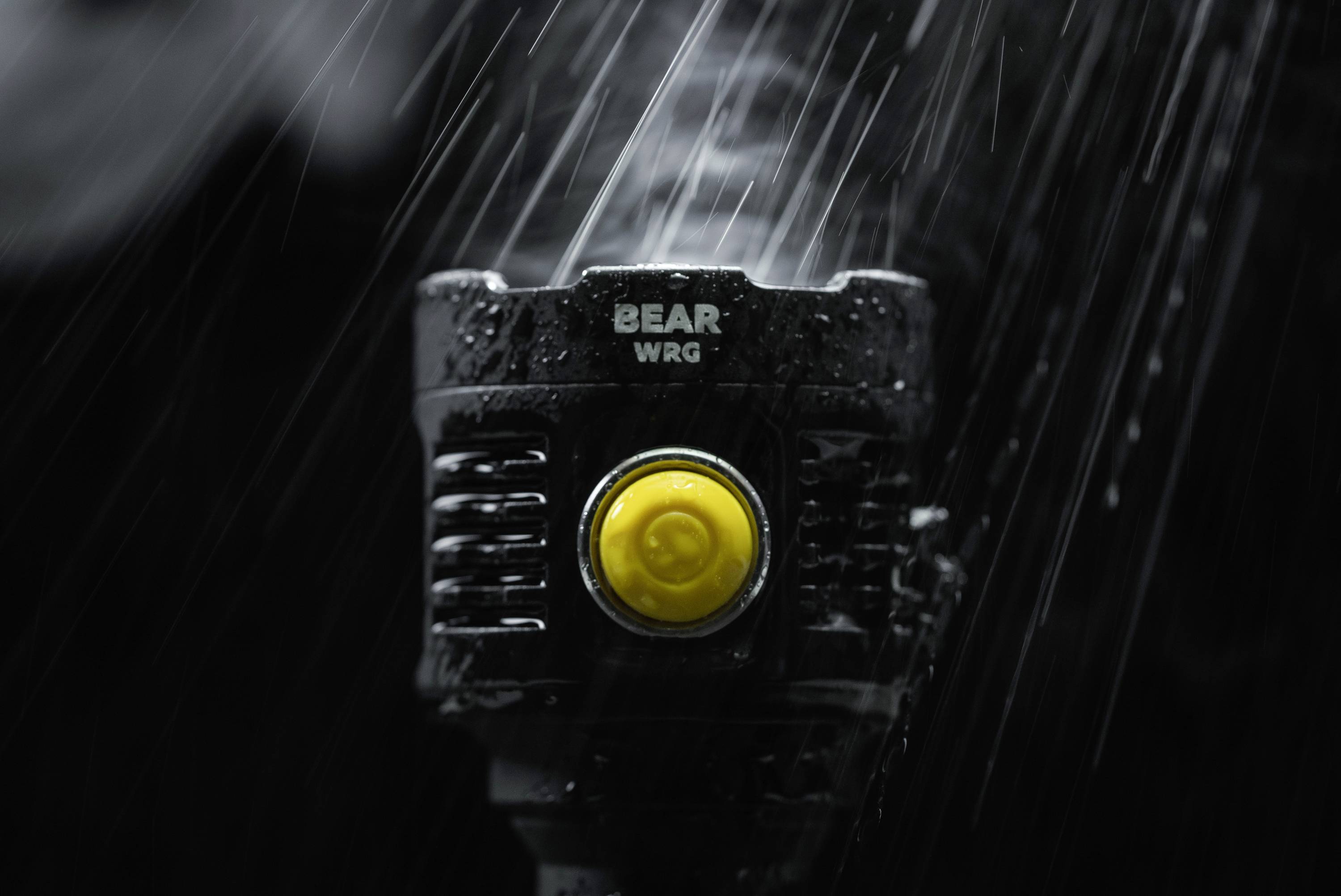 Czarne urządzenie z żółtym przyciskiem, z napisem 'BEAR WRG', sfotografowane w ponurym otoczeniu podczas deszczu.
