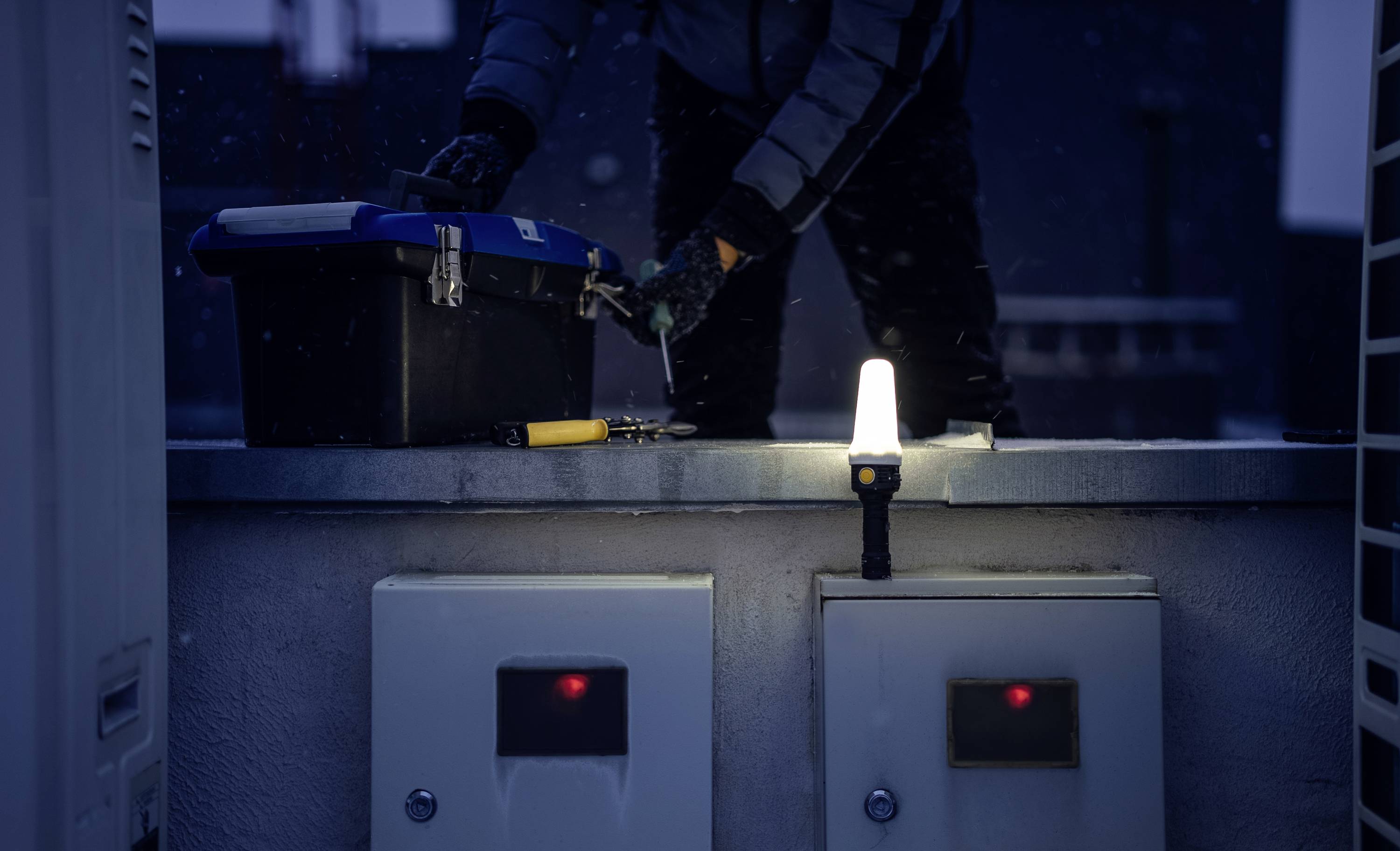 Osoba pracuje w nocy na wolnym powietrzu przy urządzeniu technicznym, oświetlona światłem latarki. Scena przedstawia narzędzia leżące na powierzchni.