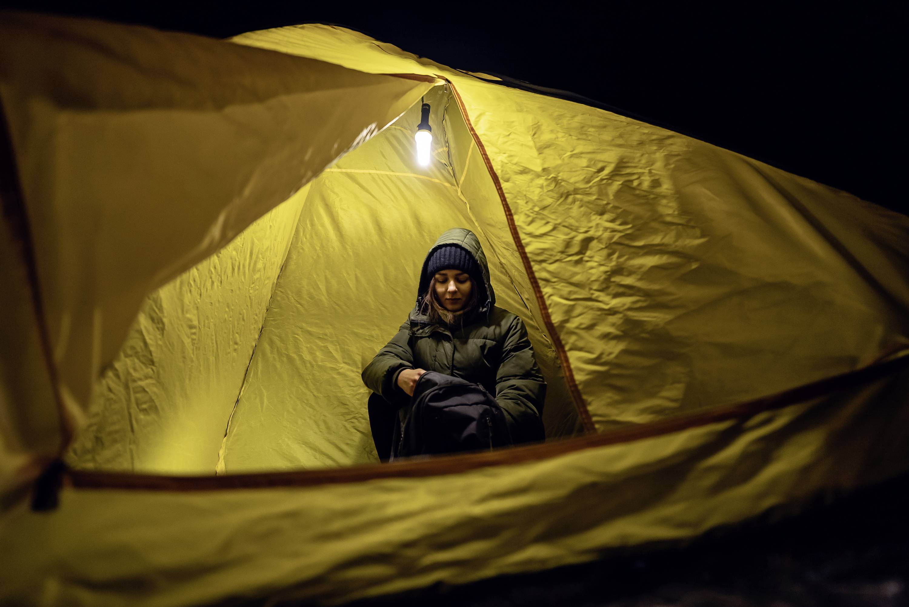 Osoba siedzi w nocy w żółtym namiocie, ubrana w ciepłą odzież, wyglądająca na rozluźnioną. Lampa oświetla wnętrze namiotu.