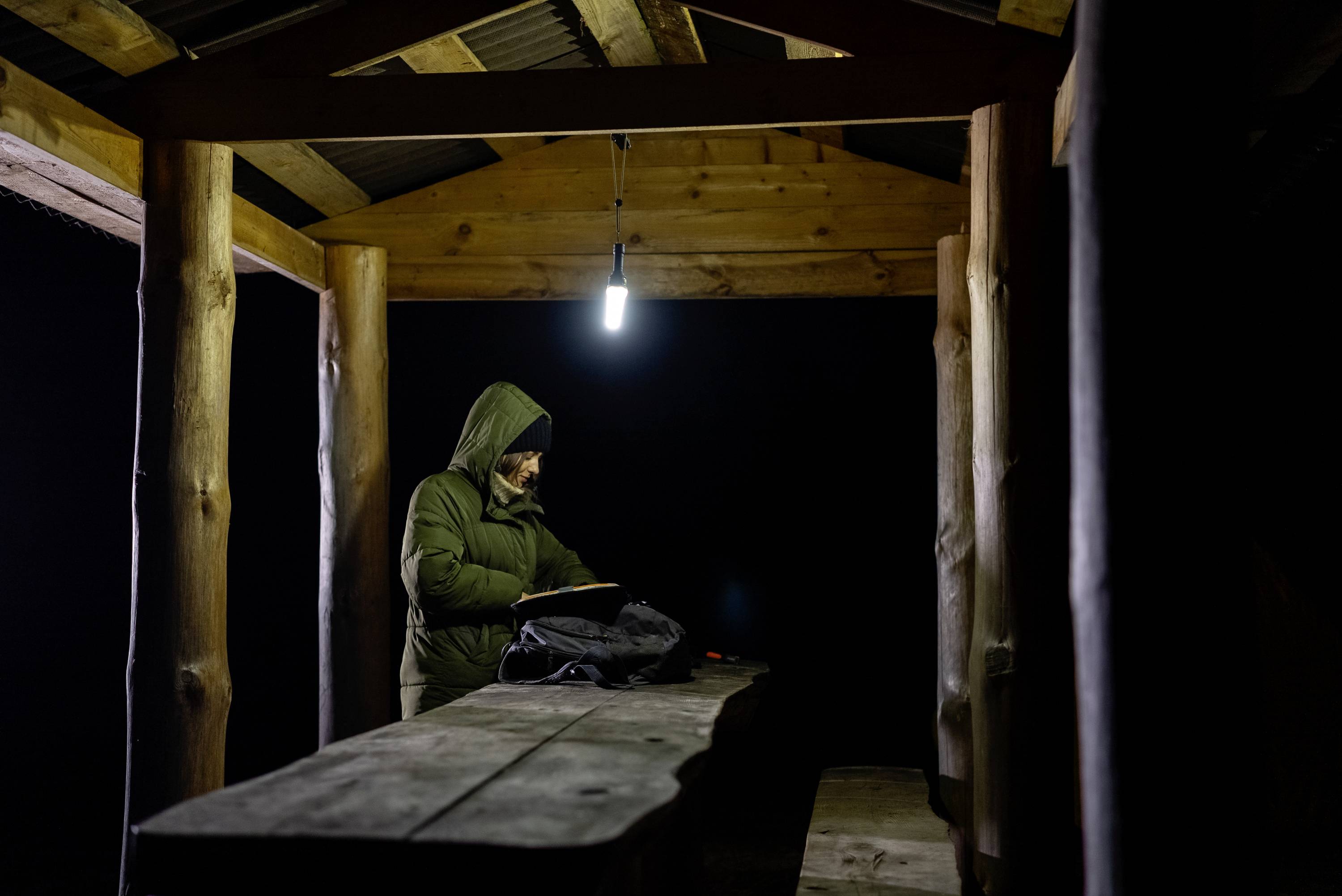 Osoba w ciepłym ubraniu stoi nocą pod oświetlonym drewnianym zadaszeniem, czyta coś i ma torbę na stole obok siebie.