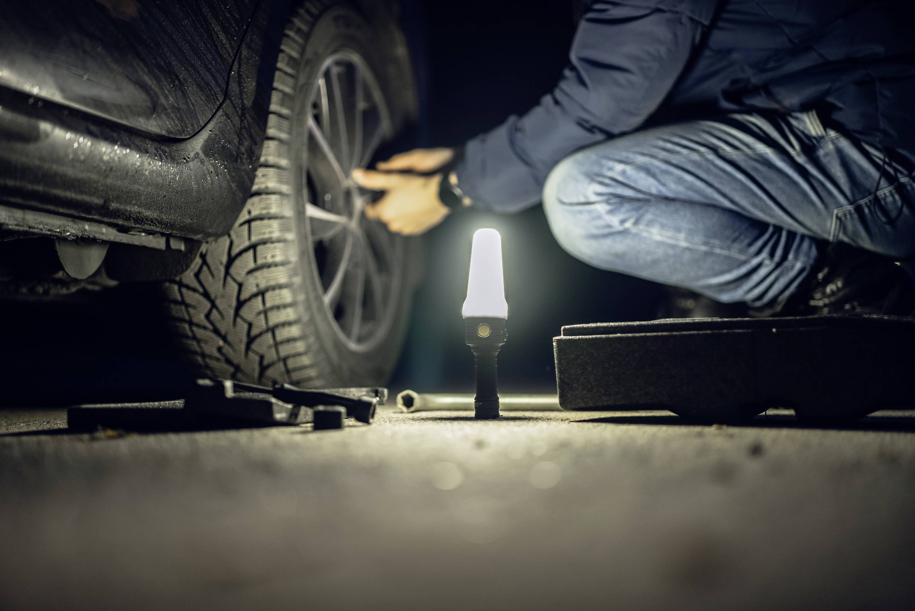 Osoba wymienia oponę w samochodzie nocą. Lampa oświetla obszar pracy. Różne narzędzia leżą na ziemi.