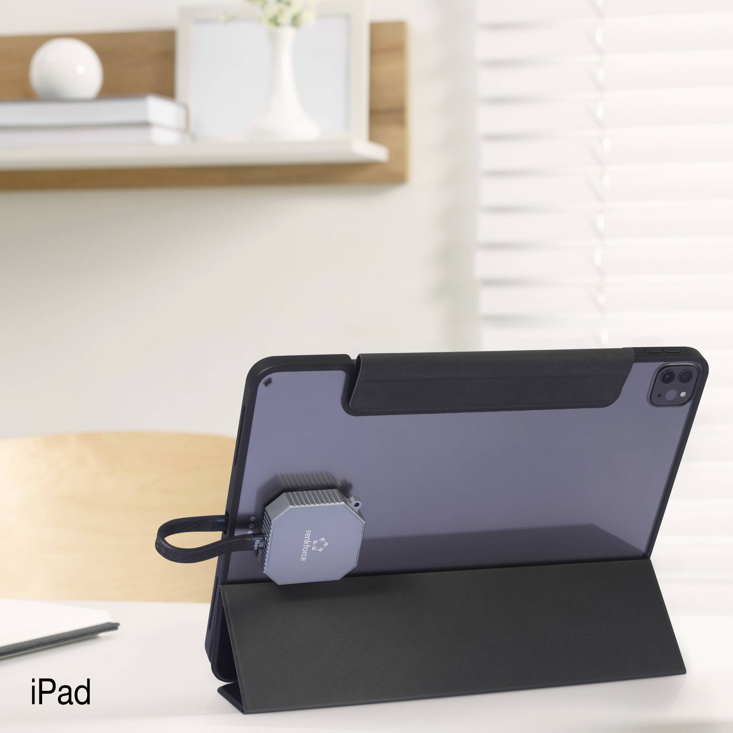 Tablet iPad w czarnym etui stoi na stole, podłączone do ładowarki. W tle widać regał z książkami.