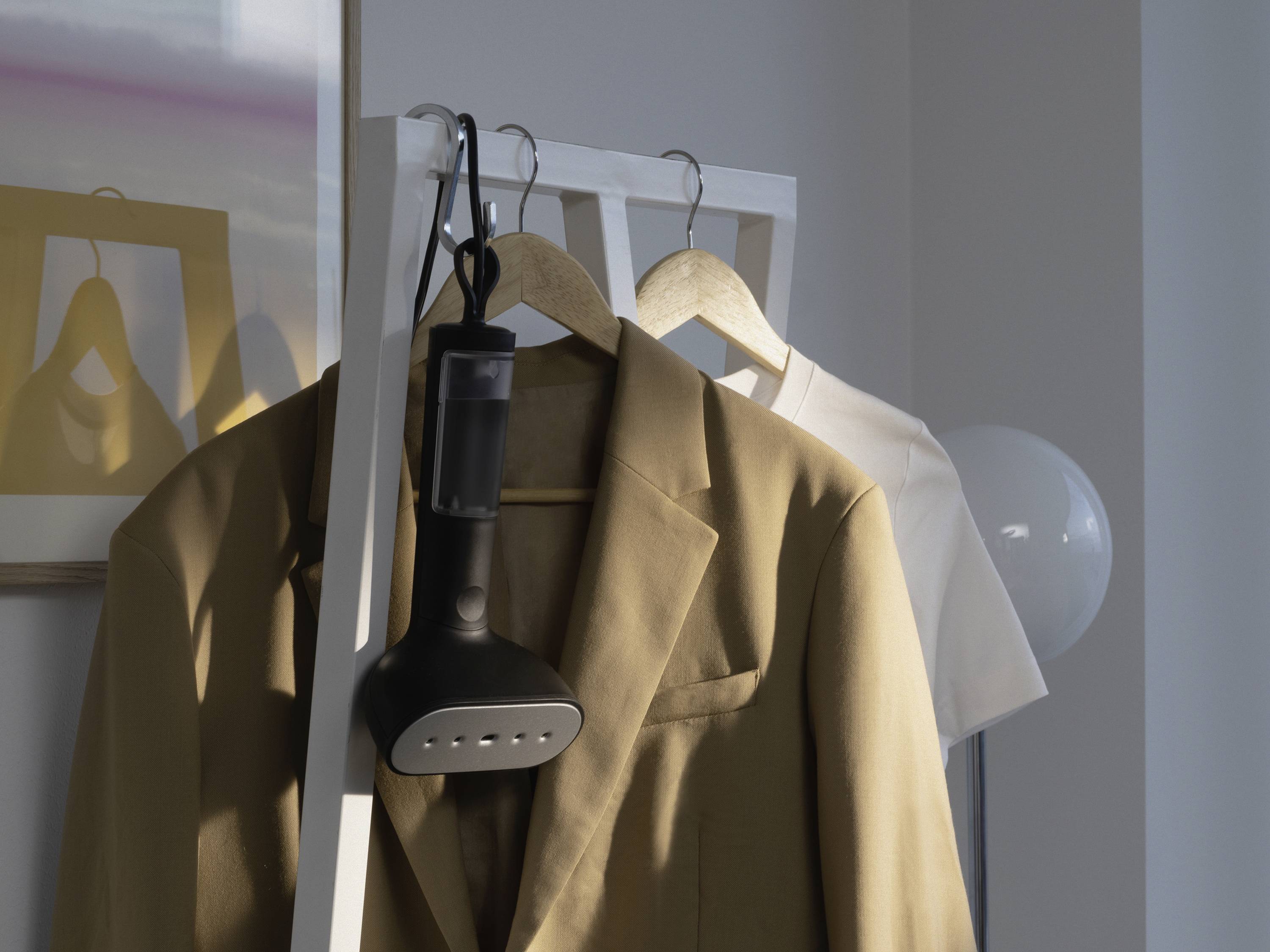 Beżowy garnitur wisi na wieszaku, obok niego biała koszula. Żelazko parowe jest zamocowane na drążku.