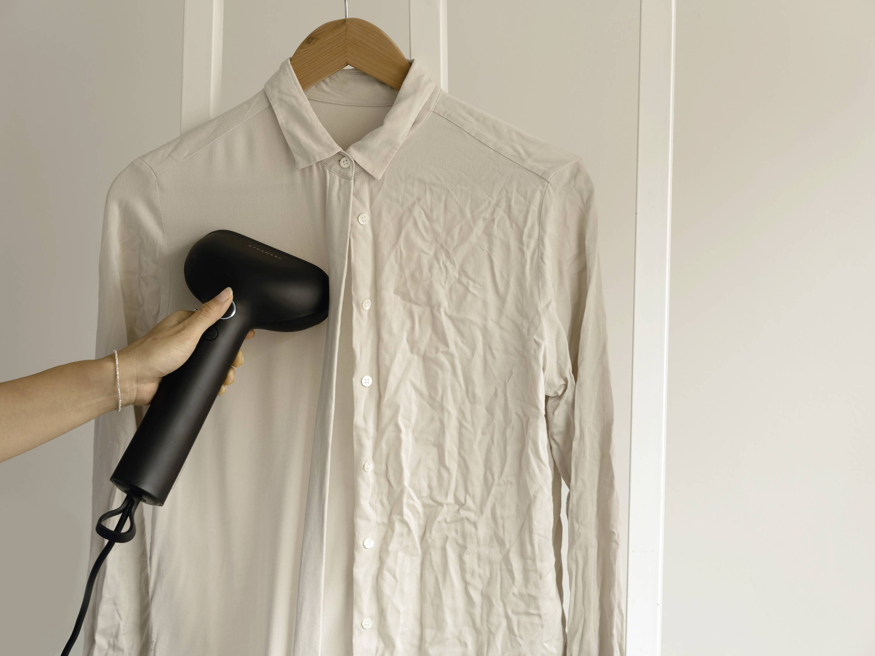 Dłoń wygładza białą koszulę na wieszaku czarnym żelazkiem parowym. Koszula naprasowana w połowie.