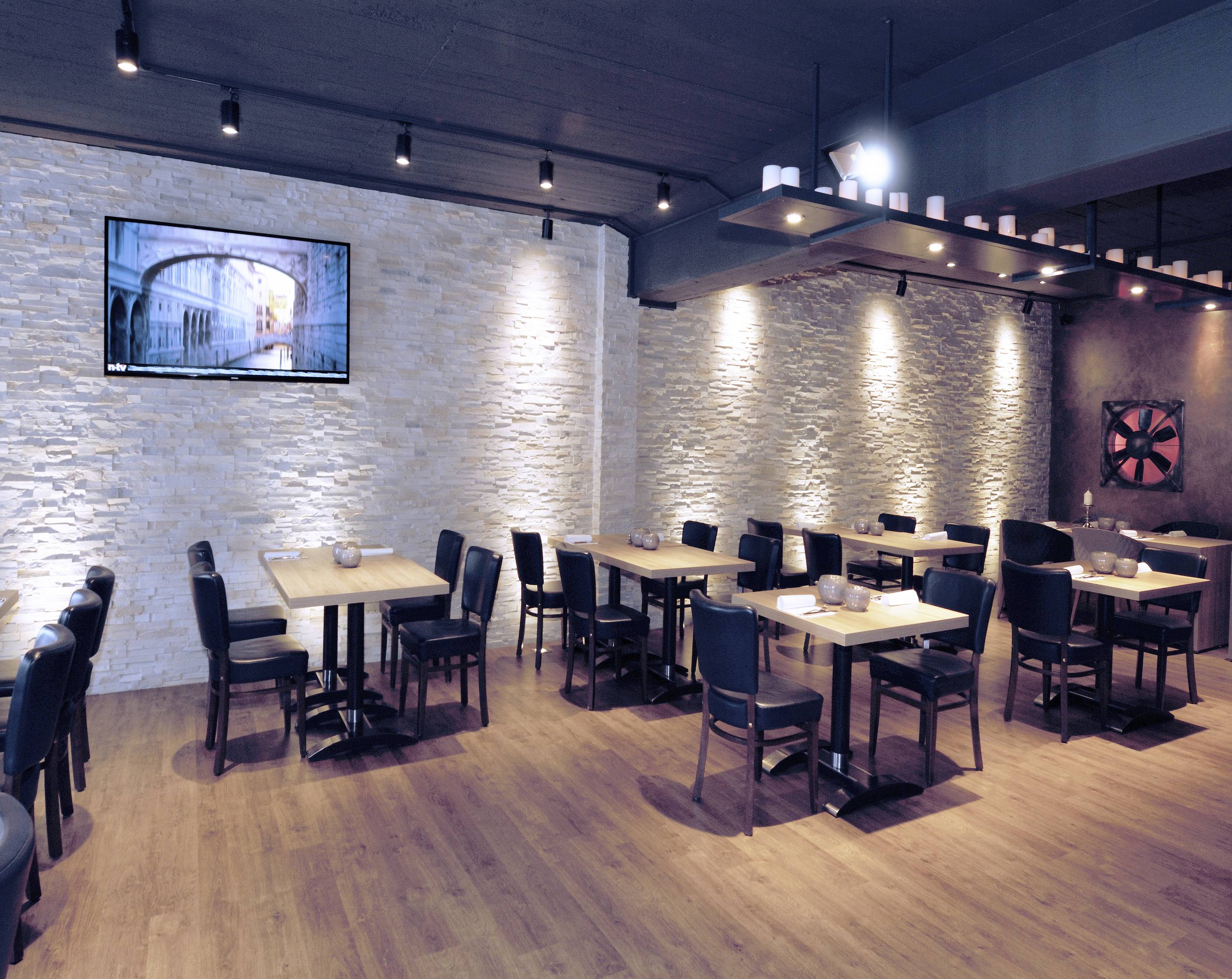 Nowoczesna restauracja z drewnianą podłogą, białymi ścianami z cegły i czarnymi krzesłami. Puste stoły ustawione są w rzędach, a na ścianie wisi ekran.