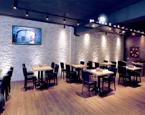 Nowoczesna restauracja z drewnianą podłogą, białymi ścianami z cegły i płaskim telewizorem zamontowanym na ścianie. Stoły i krzesła są równomiernie rozmieszczone.