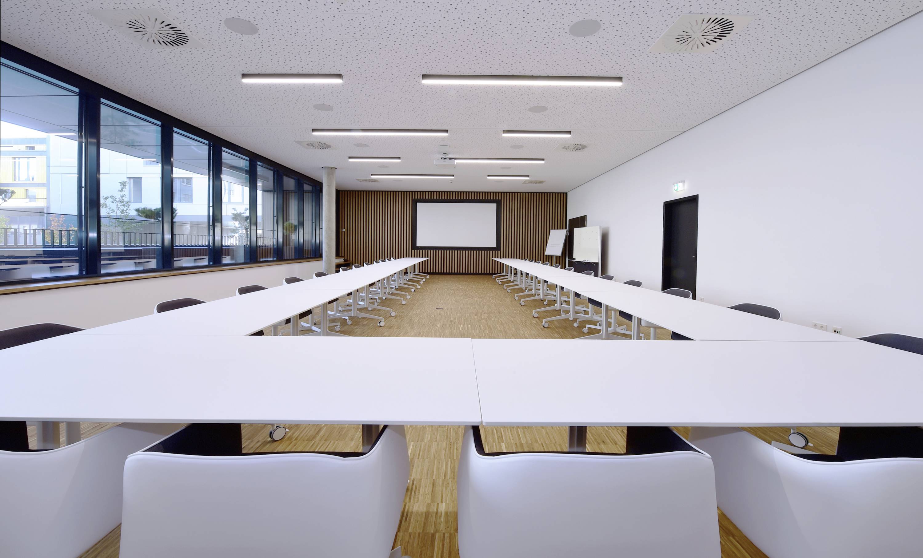 Nowoczesna sala konferencyjna z ustawionymi w kształcie litery U stołami, białymi krzesłami, dużymi oknami oraz ekranem umieszczonym z przodu przy ścianie.