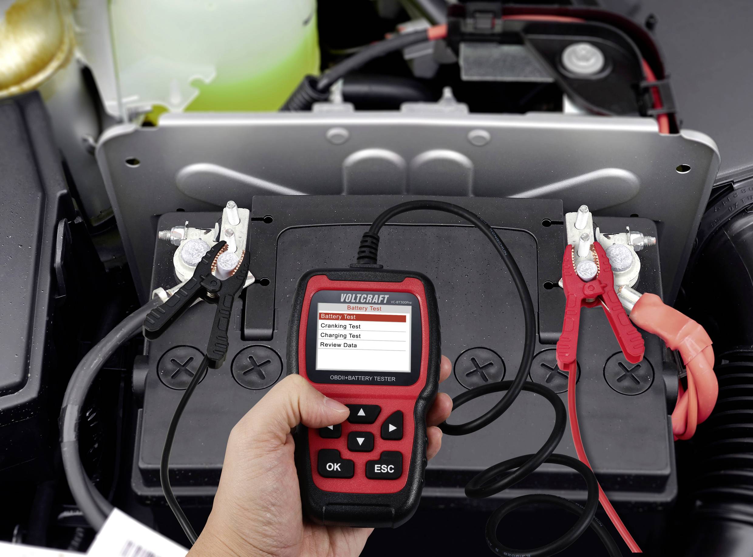 Dłoń trzyma urządzenie do testowania akumulatora podłączone do akumulatora samochodowego. Na wyświetlaczu widoczne są różne opcje testowania.