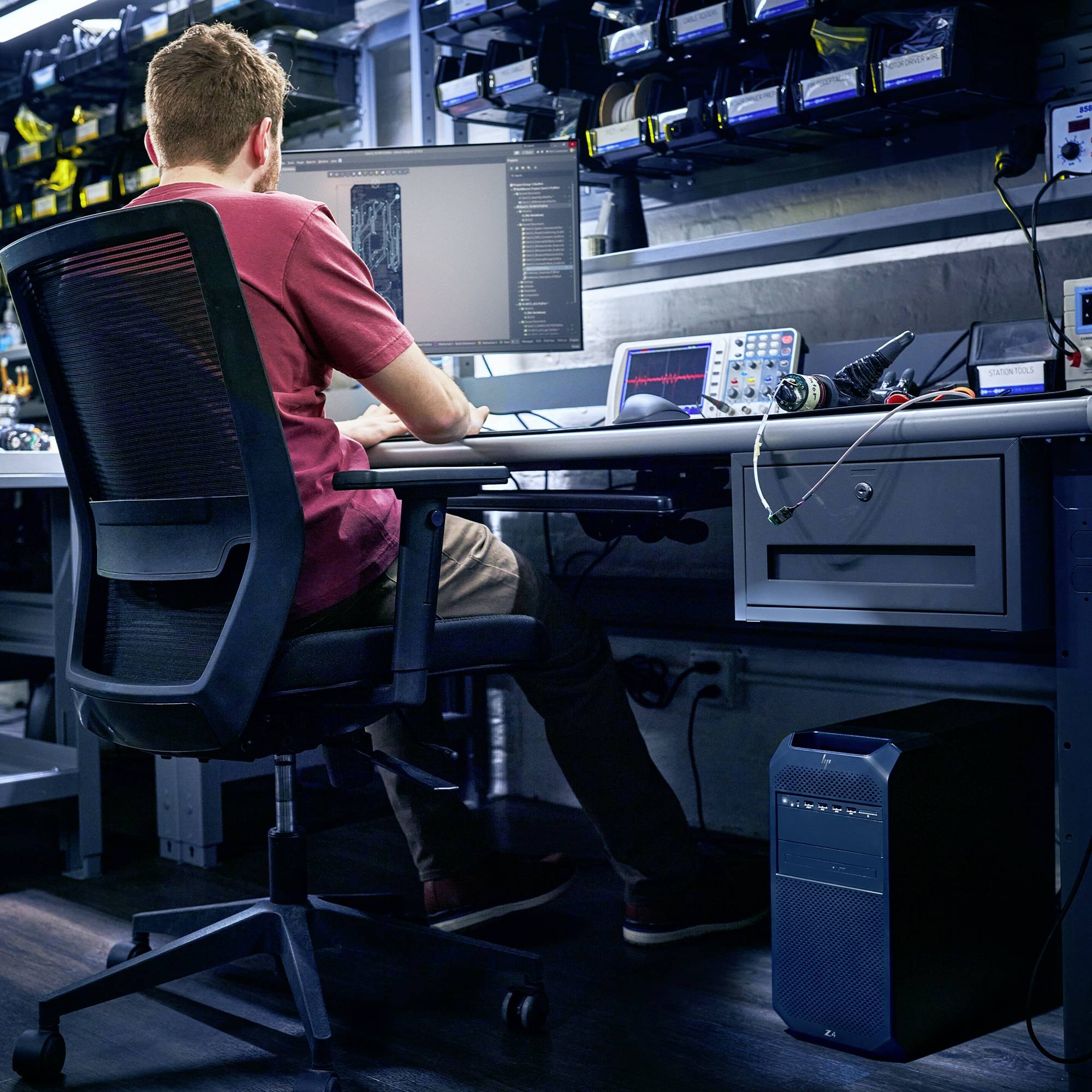 Osoba siedzi przy biurku i pracuje na komputerze z wieloma monitorami w technologicznie wyposażonym pomieszczeniu.