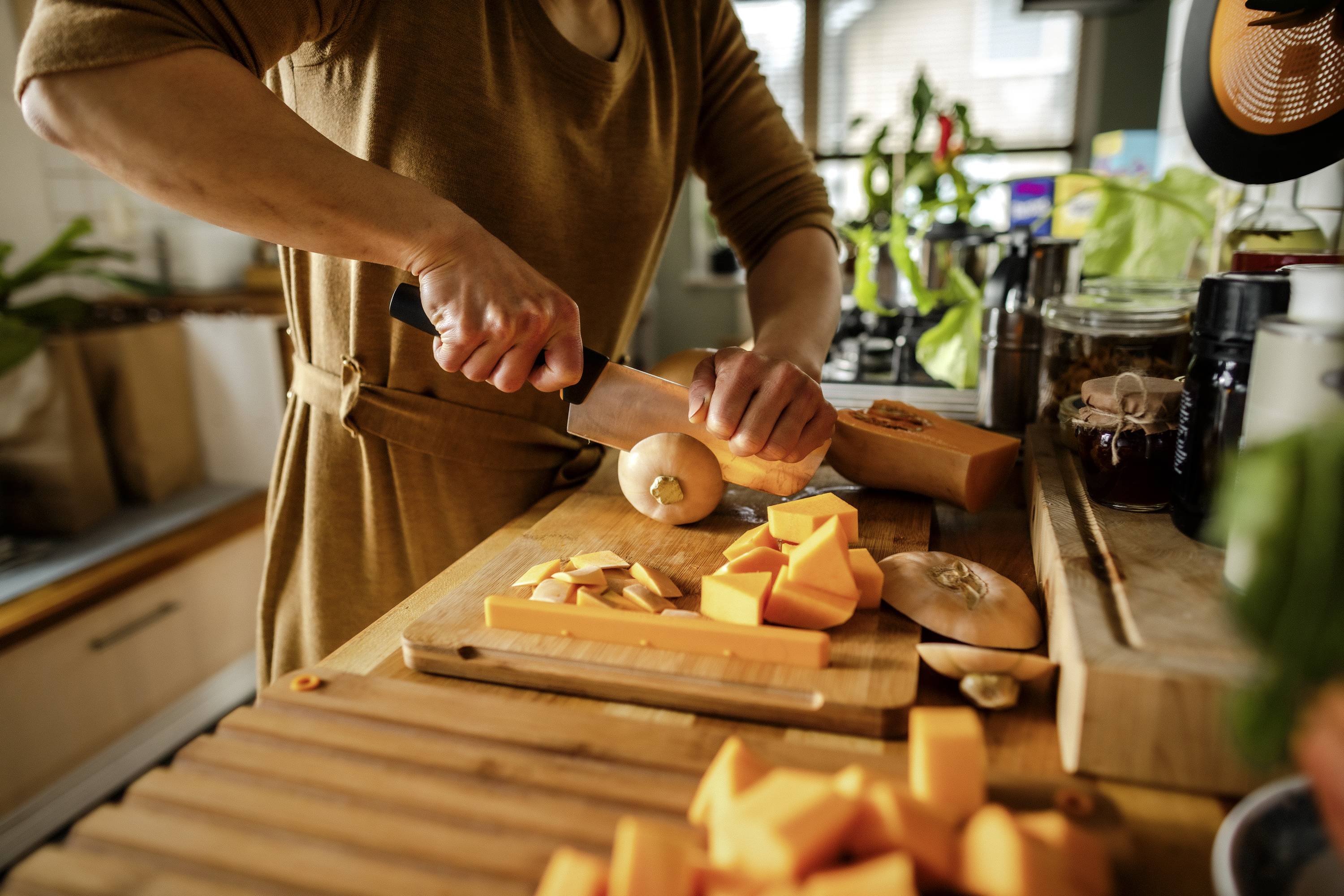Osoba kroi dynię na małe kostki na drewnianej desce w kuchni. W tle widać różne przybory kuchenne.