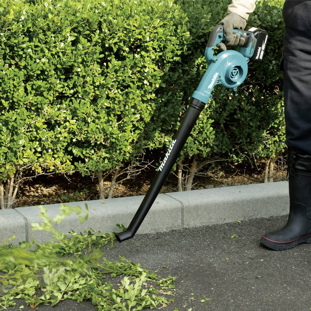 Osoba używa turkusowego odkurzacza do liści, aby oczyścić zielone liście ze ścieżki ogrodowej, z przystrzyżonymi krzewami w tle.