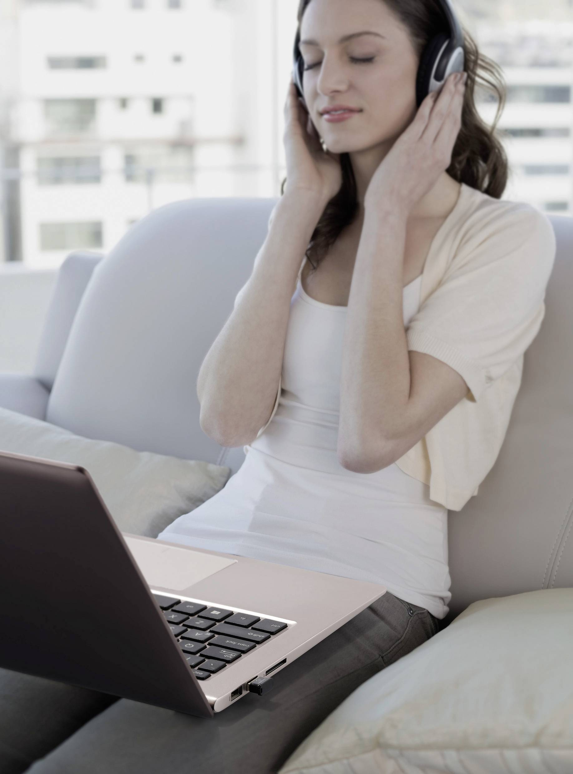 Kobieta z zamkniętymi oczami słucha muzyki przez słuchawki i siedzi zrelaksowana na kanapie z laptopem na kolanach.