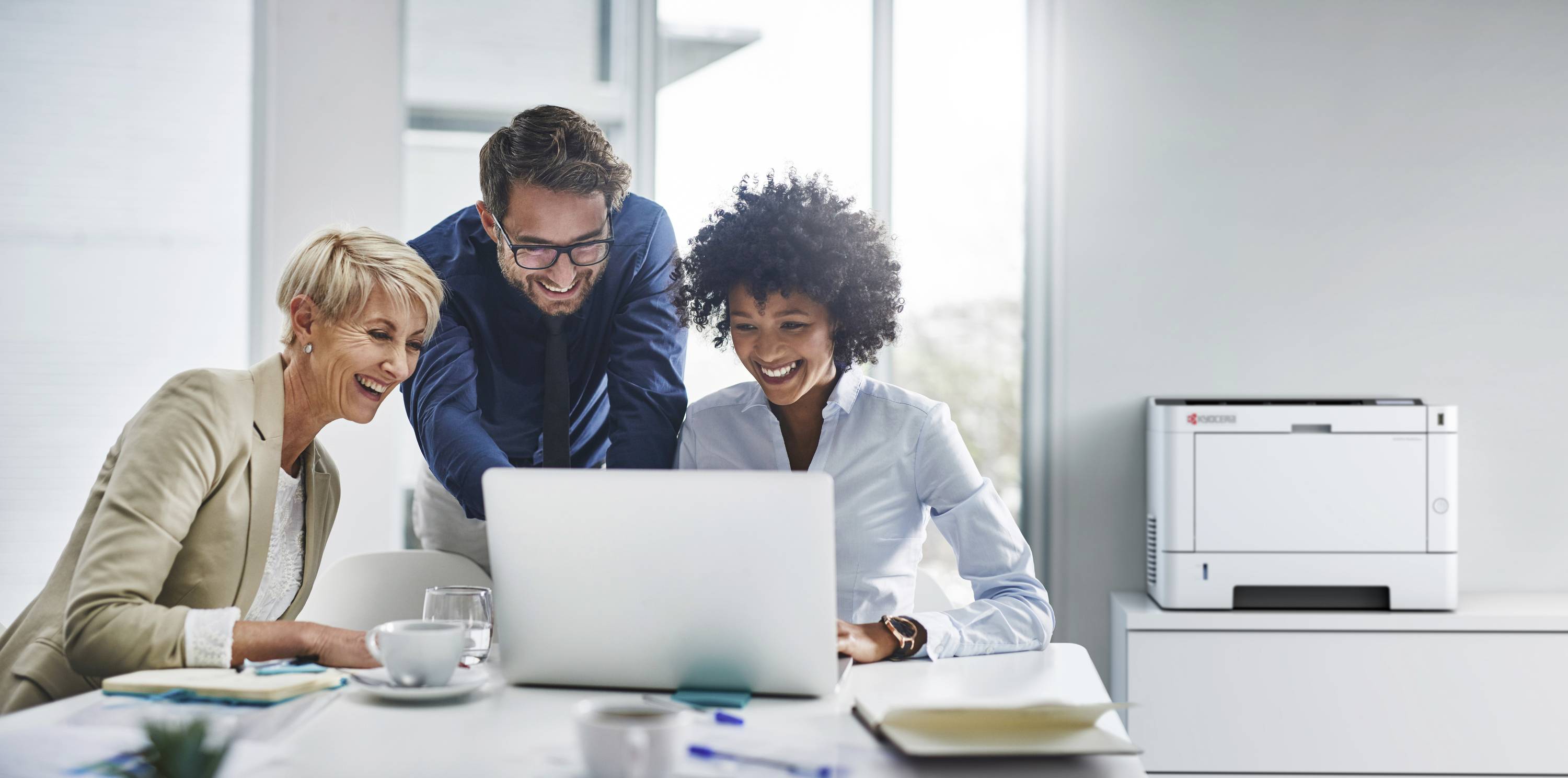 Trzech osób uśmiecha się i współpracuje przy laptopie w jasnym biurze, drukarka stoi w tle na stole.
