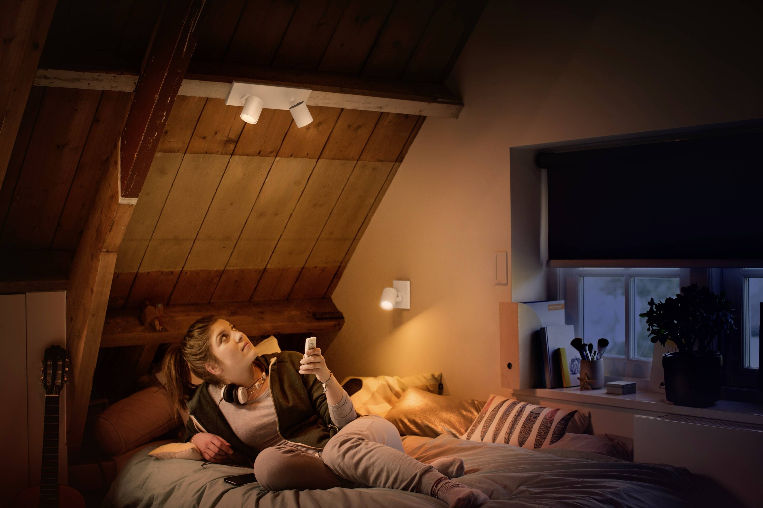 Kobieta leży na łóżku w przytulnie oświetlonym poddaszu i trzyma pilota, patrząc w górę.