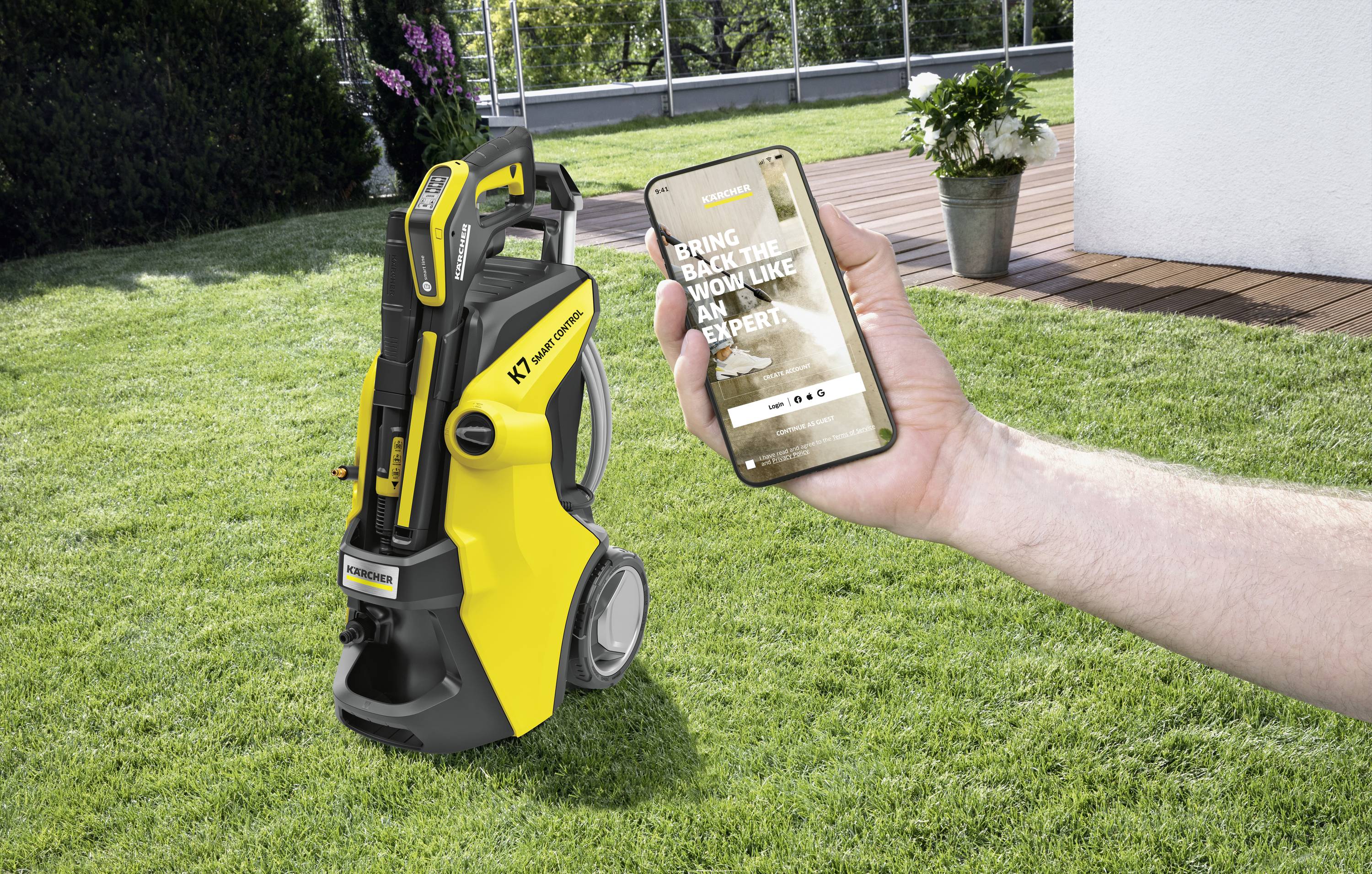 Myjka ciśnieniowa stoi na trawniku. Ręka trzyma smartfon wyświetlający aplikację do obsługi myjki ciśnieniowej.