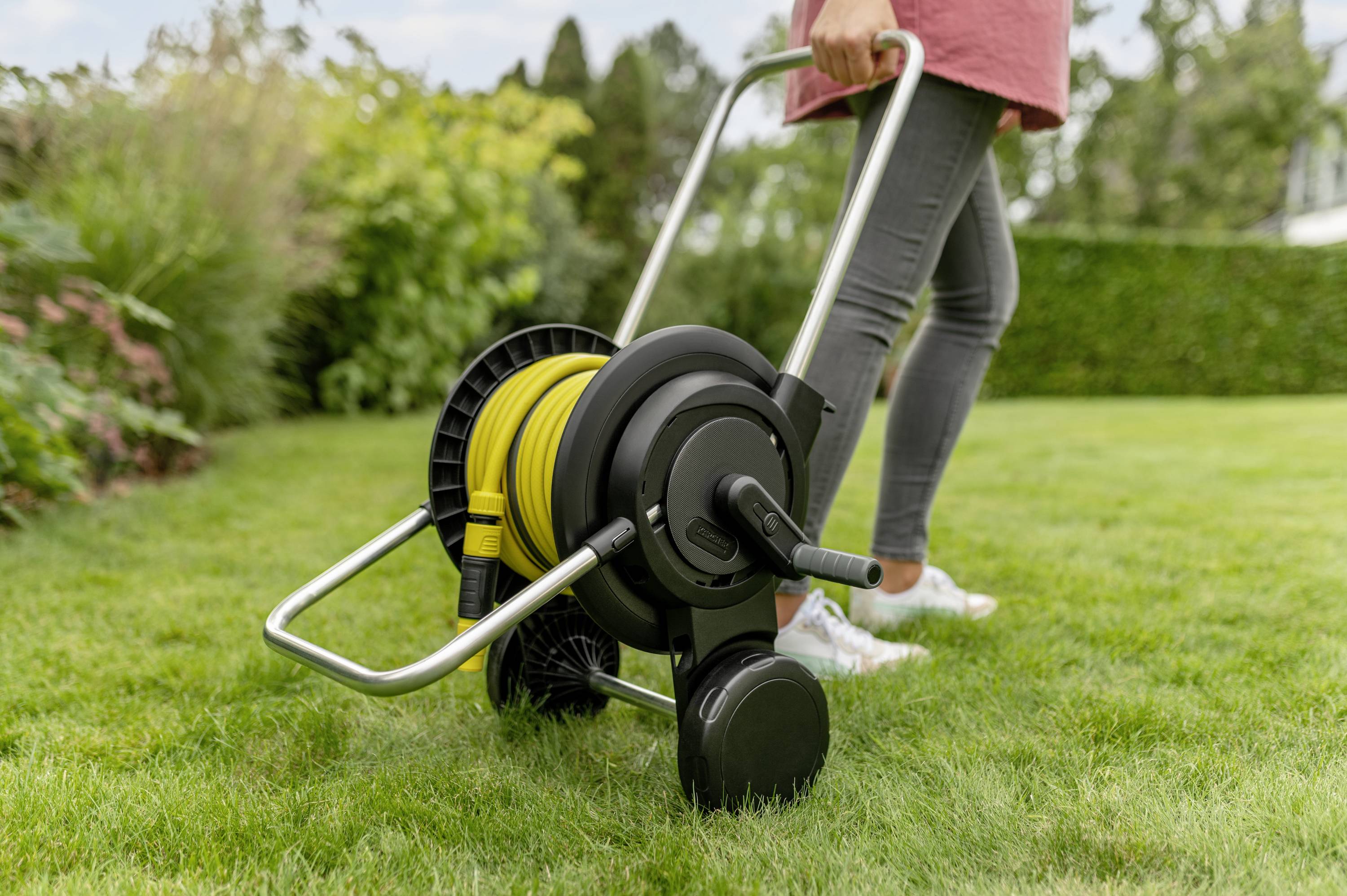 Osoba ciągnie mobilny wózek na wąż ogrodowy przez trawnik. Wąż jest żółty, a wózek posiada metalową ramę.