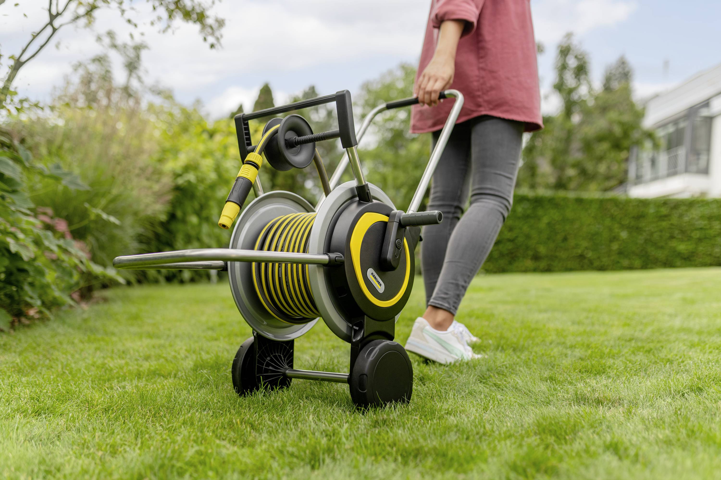 Osoba ciągnie wózek na wąż ogrodowy z żółto-czarnym wężem przez zielony trawnik w ogrodzie.