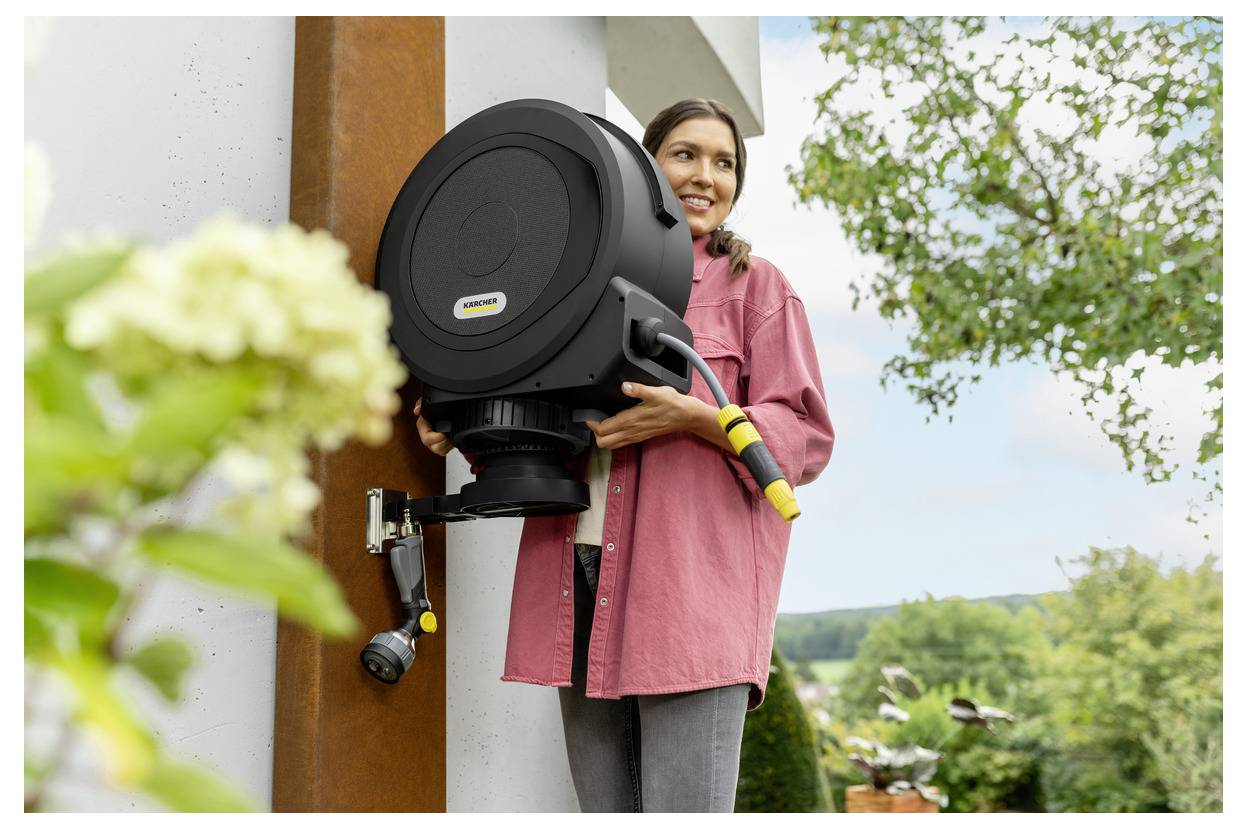 Osoba ubrana w różową koszulkę instaluje czarny zwijacz węża ogrodowego na ścianie na zewnątrz. W tle widoczne są kwitnąca roślina i drzewa.