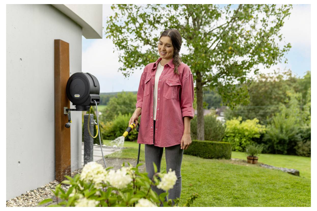 Osoba podlewa rośliny w ogrodzie za pomocą węża, otoczona zielenią i drzewem, w słoneczny dzień.