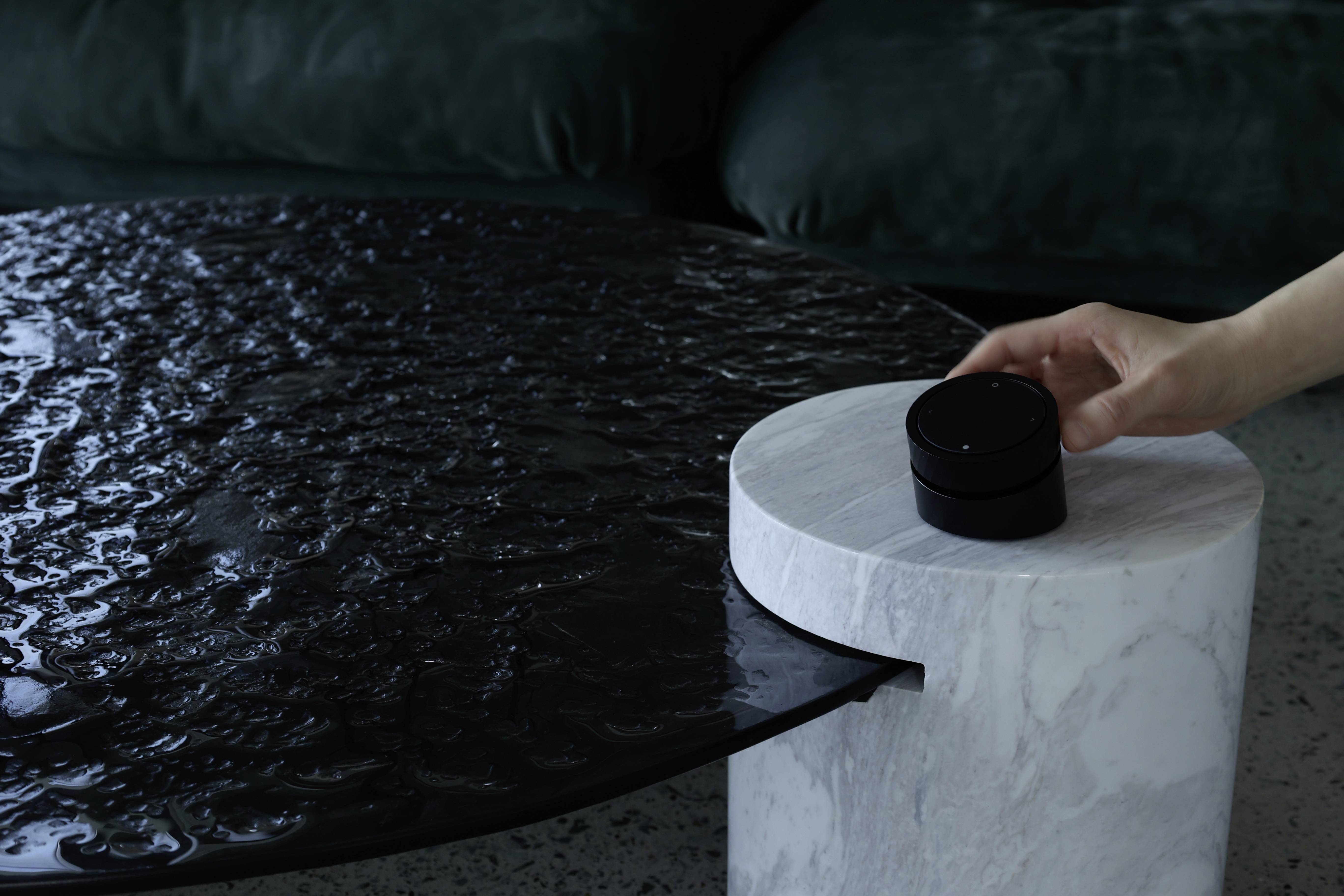 Dłoń sięga po czarny, okrągły przedmiot na białym, przypominającym marmur postumencie, obok ciemnej powierzchni stołu.