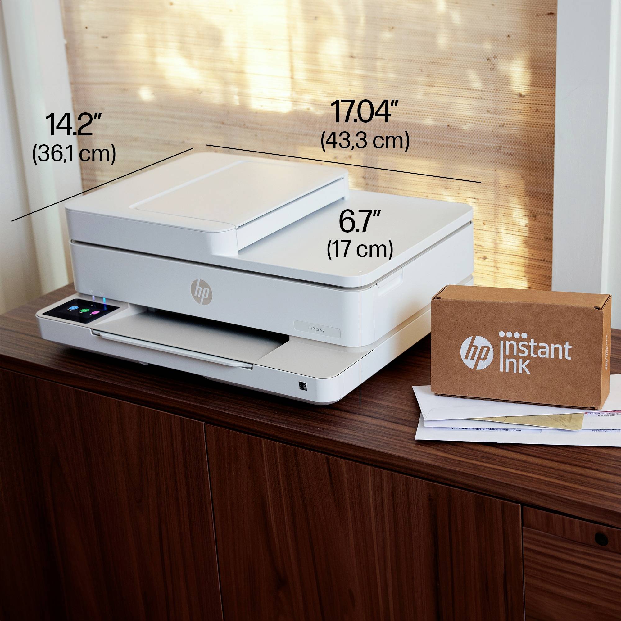 Drukarka stoi na drewnianym stole. Obok niej leży pudełko z nadrukiem 'HP Instant Ink'. Podane są wymiary drukarki.