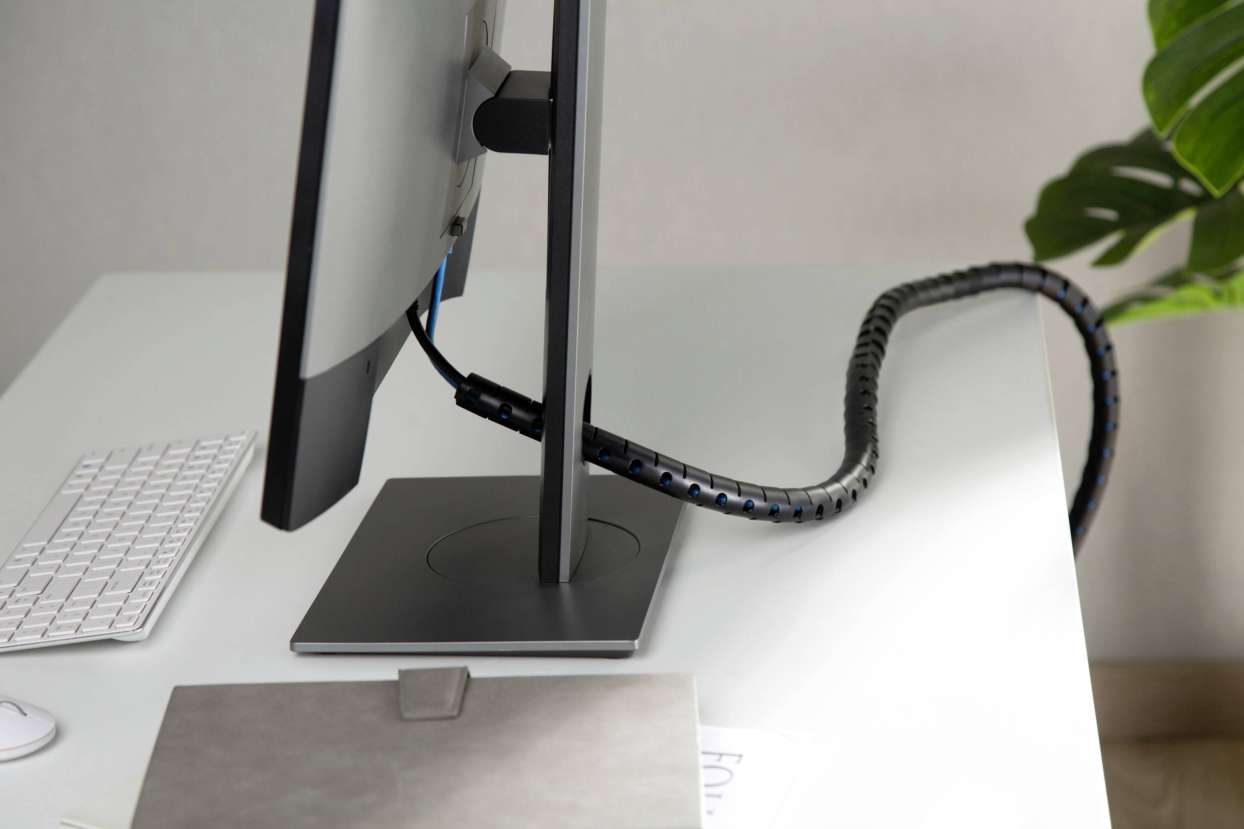 Monitor na biurku z uporządkowanymi kablami. Obok leży biała klawiatura i laptop.