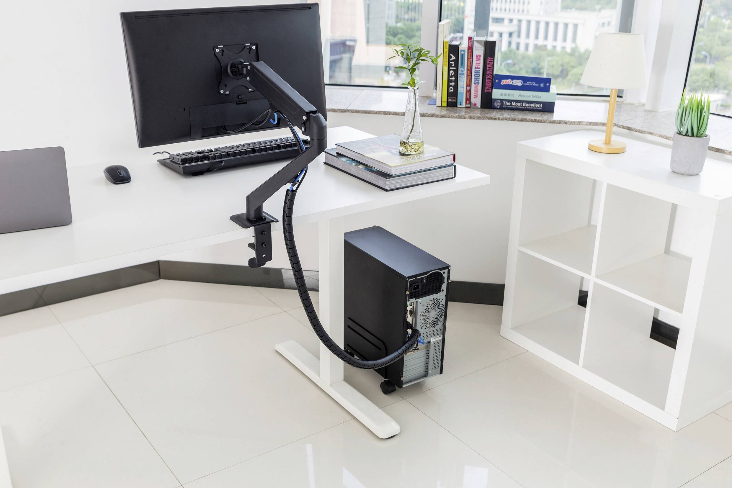 Biuro z białym stołem, na którym stoi monitor na ramieniu obrotowym. Pod spodem czarny komputer. W tle okno z widokiem na miasto.