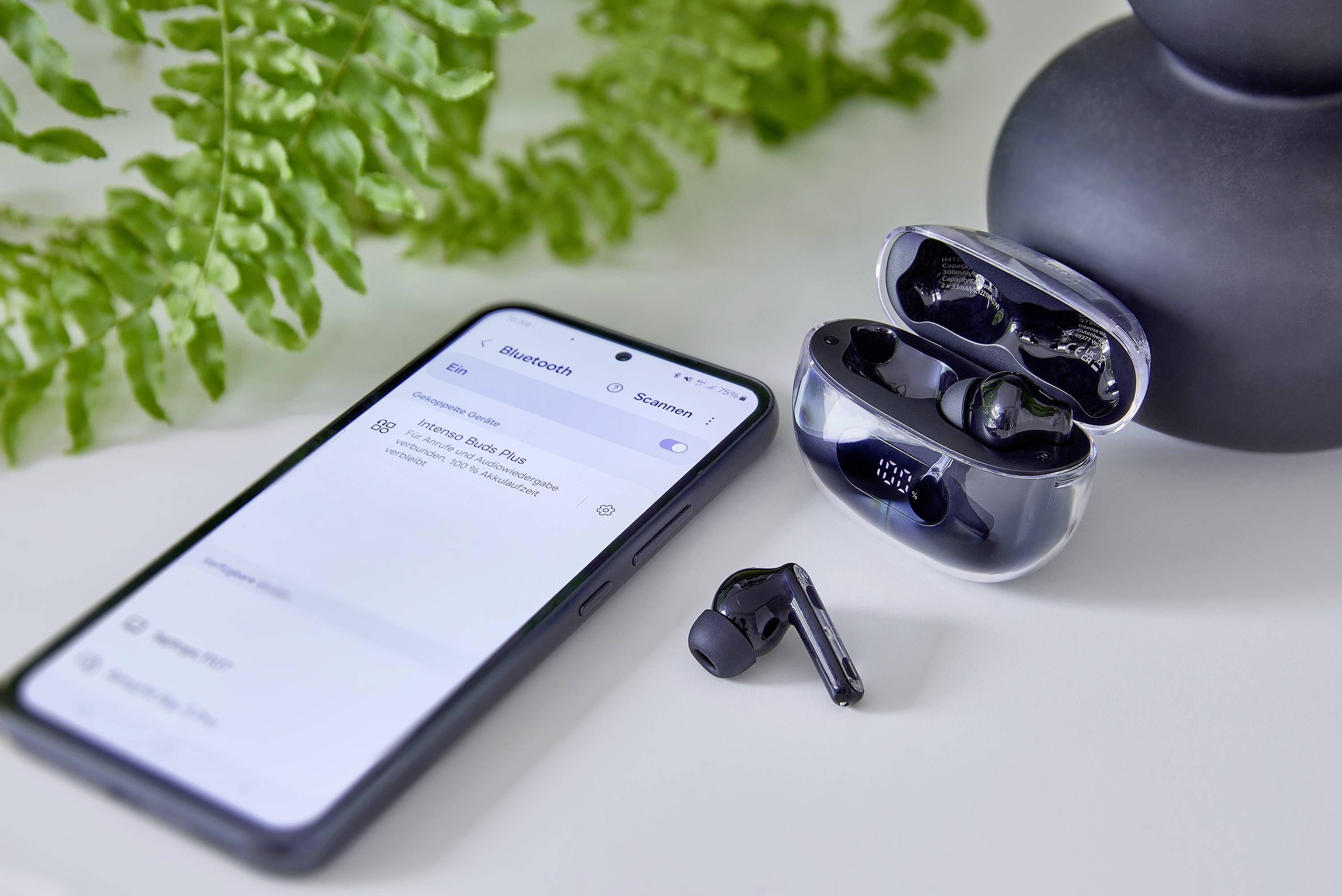 Smartfon wyświetlający ustawienia Bluetooth obok bezprzewodowych słuchawek dousznych w otwartym etui ładującym. Na tle widoczna jest roślina.