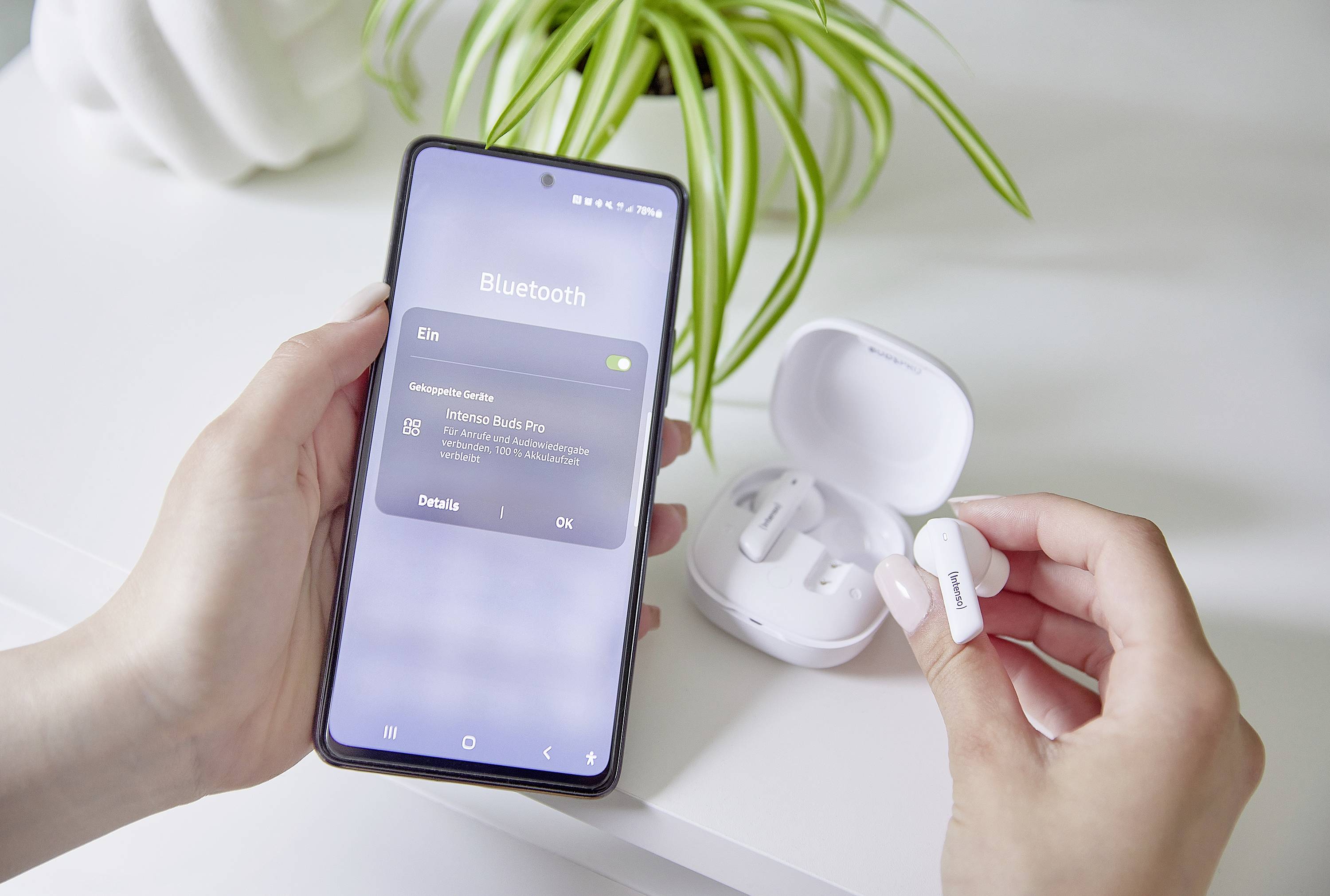 Osoba trzyma smartfon z włączonym Bluetooth, pokazując połączenie z 'Hama Buds Pro'. Lewa ręka trzyma słuchawkę douszną. W tle widoczna jest roślina.