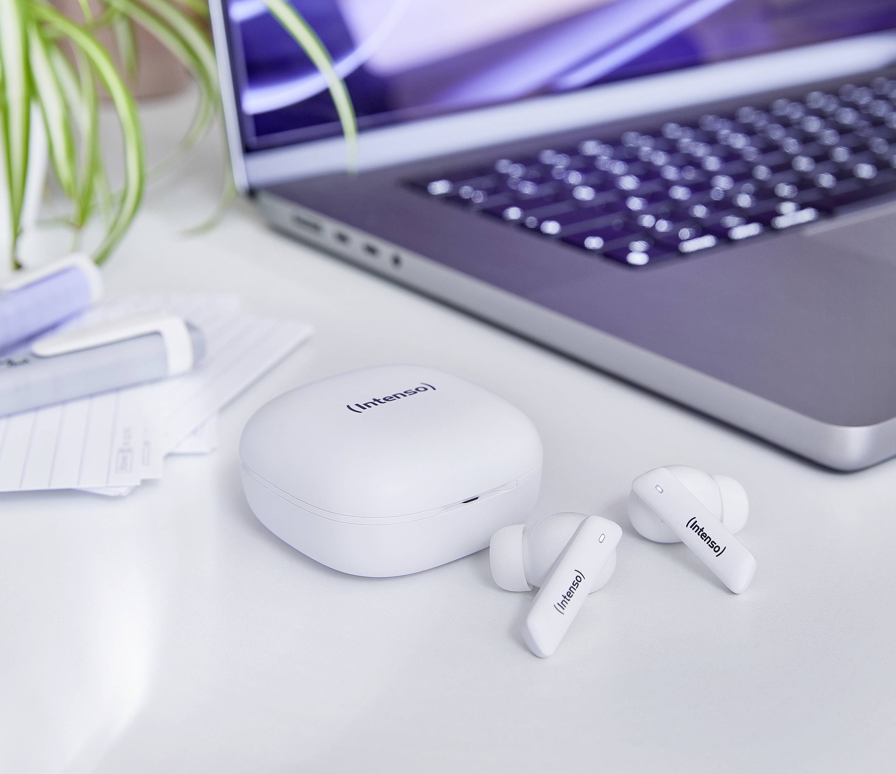 Białe bezprzewodowe słuchawki douszne i etui ładujące na białym biurku obok otwartego laptopa. W tle roślina.