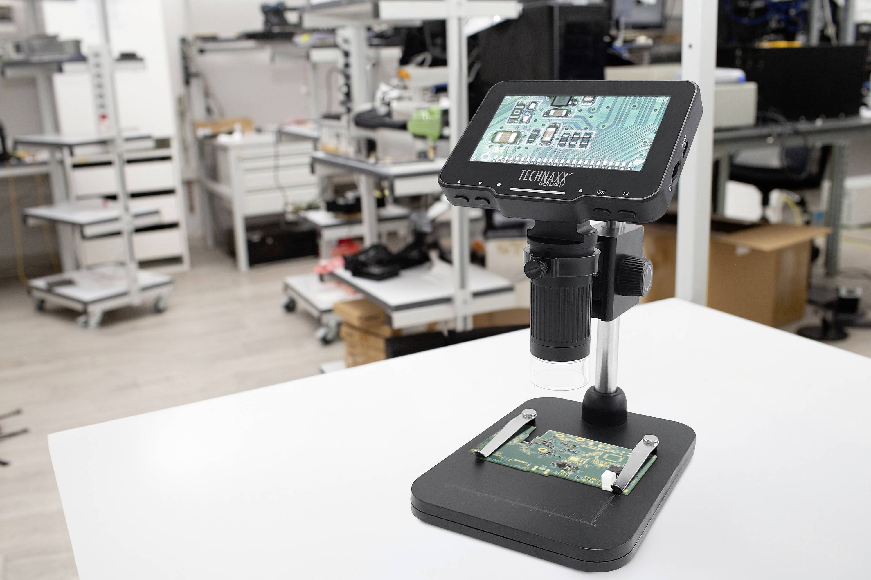 Cyfrowy mikroskop na stole w laboratorium technicznym, z płytką obwodu drukowanego pod obiektywem. W tle widoczne są różne stanowiska robocze.