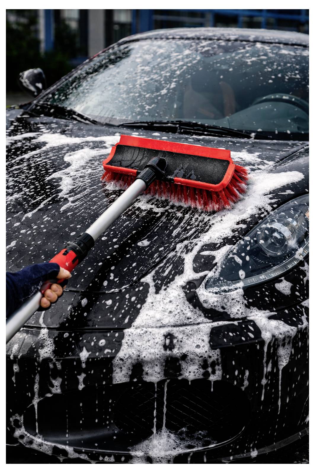 Osoba myąca czarny samochód czerwoną szczotką, pokryta pianą mydlaną.