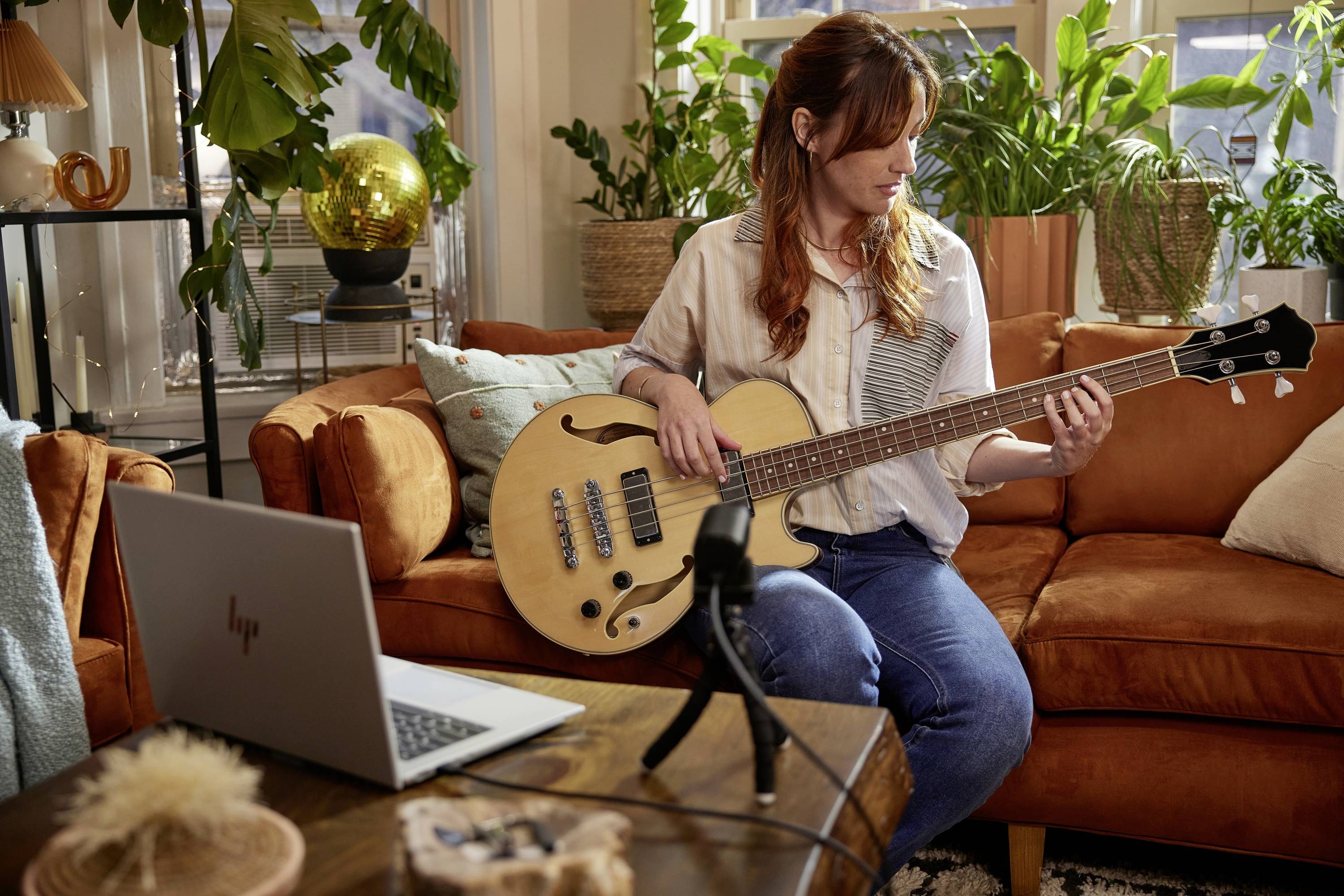 Kobieta gra na gitarze basowej siedząc na kanapie w przytulnym salonie. Przed nią na stole stoi laptop i mikrofon. W tle widać rośliny i dekoracje.