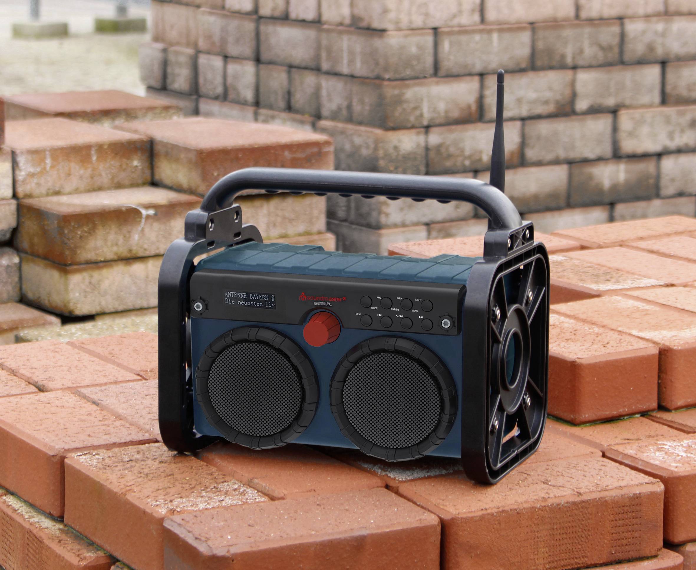Przenośne radio budowlane z dwoma głośnikami stoi na ułożonych jeden na drugim cegłach. Posiada uchwyt i antenę.