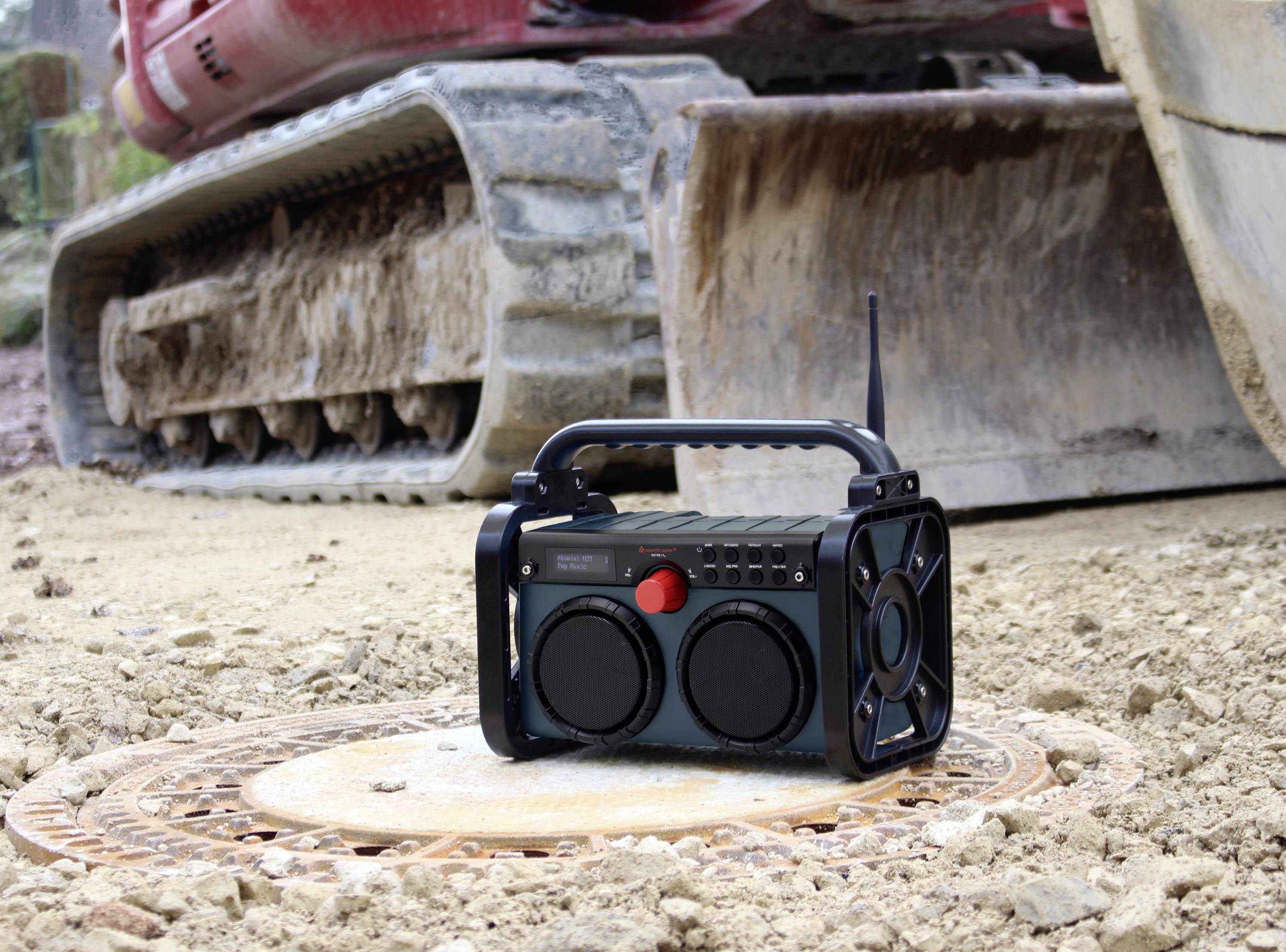 Przenośne radio stoi na pokrywie włazu kanalizacyjnego na placu budowy, w tle widać koparkę.