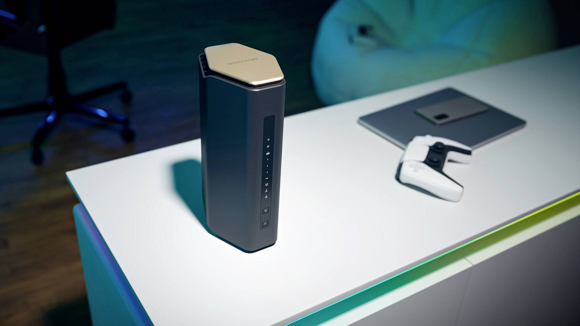 Czarny router Wi-Fi na biurku obok kontrolera i laptopa. W tle fotel biurowy i worek sako w słabo oświetlonym pomieszczeniu.