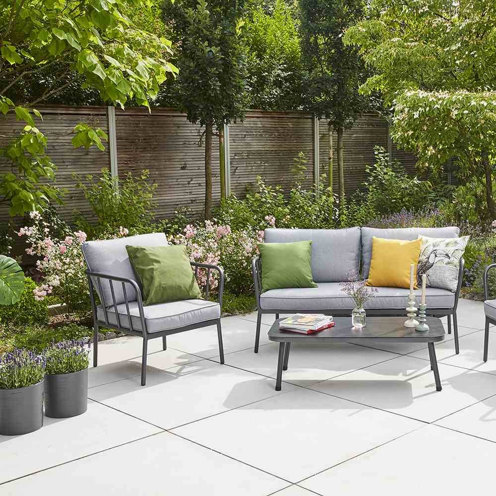Zestaw mebli ogrodowych z szarymi sofami i zielonymi poduszkami na tarasie, otoczony kwitnącymi roślinami i drzewami w tle.